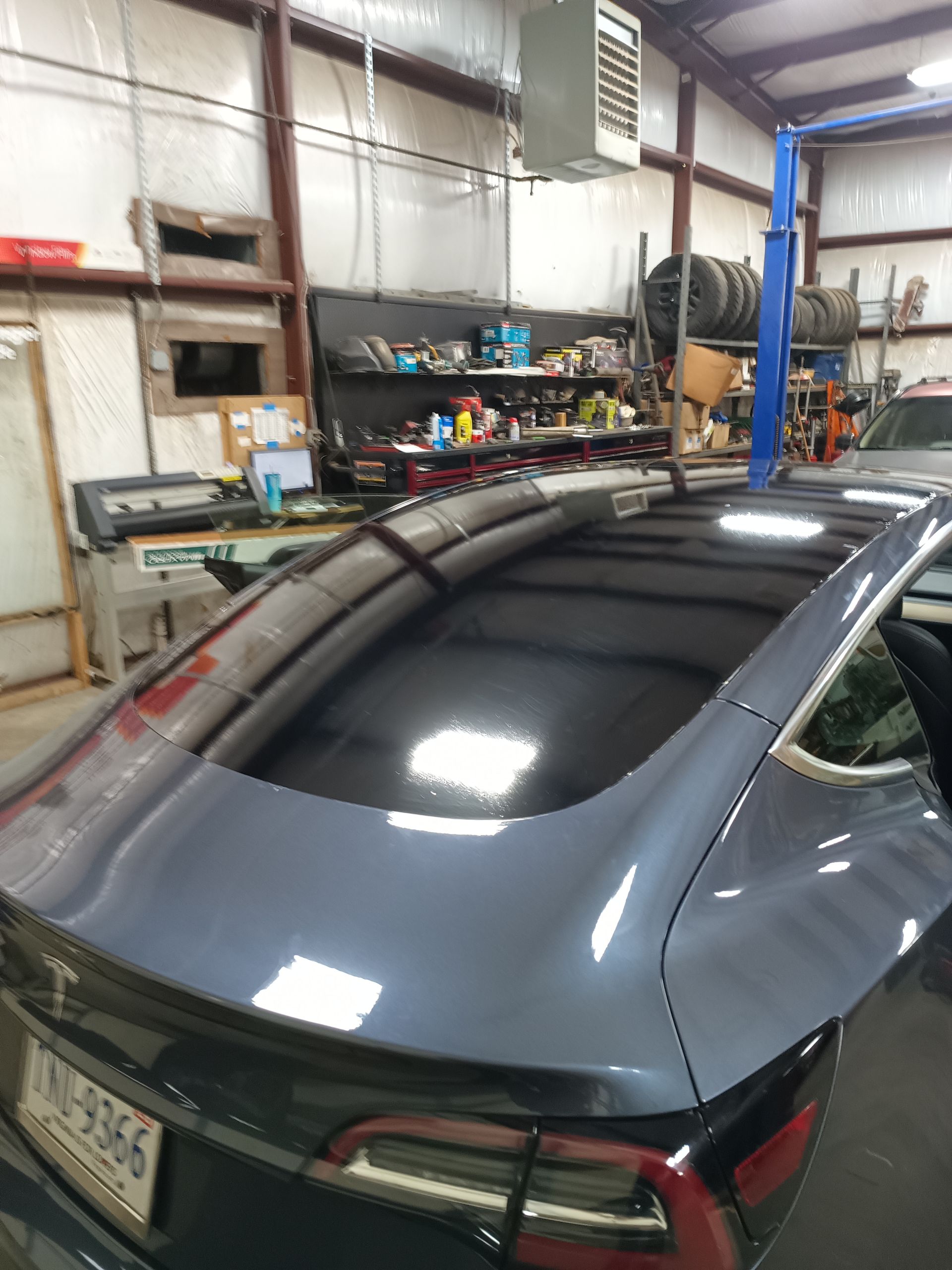 Dark gray Tesla car in a garage, shiny black roof, red tail light, shop tools in the background.