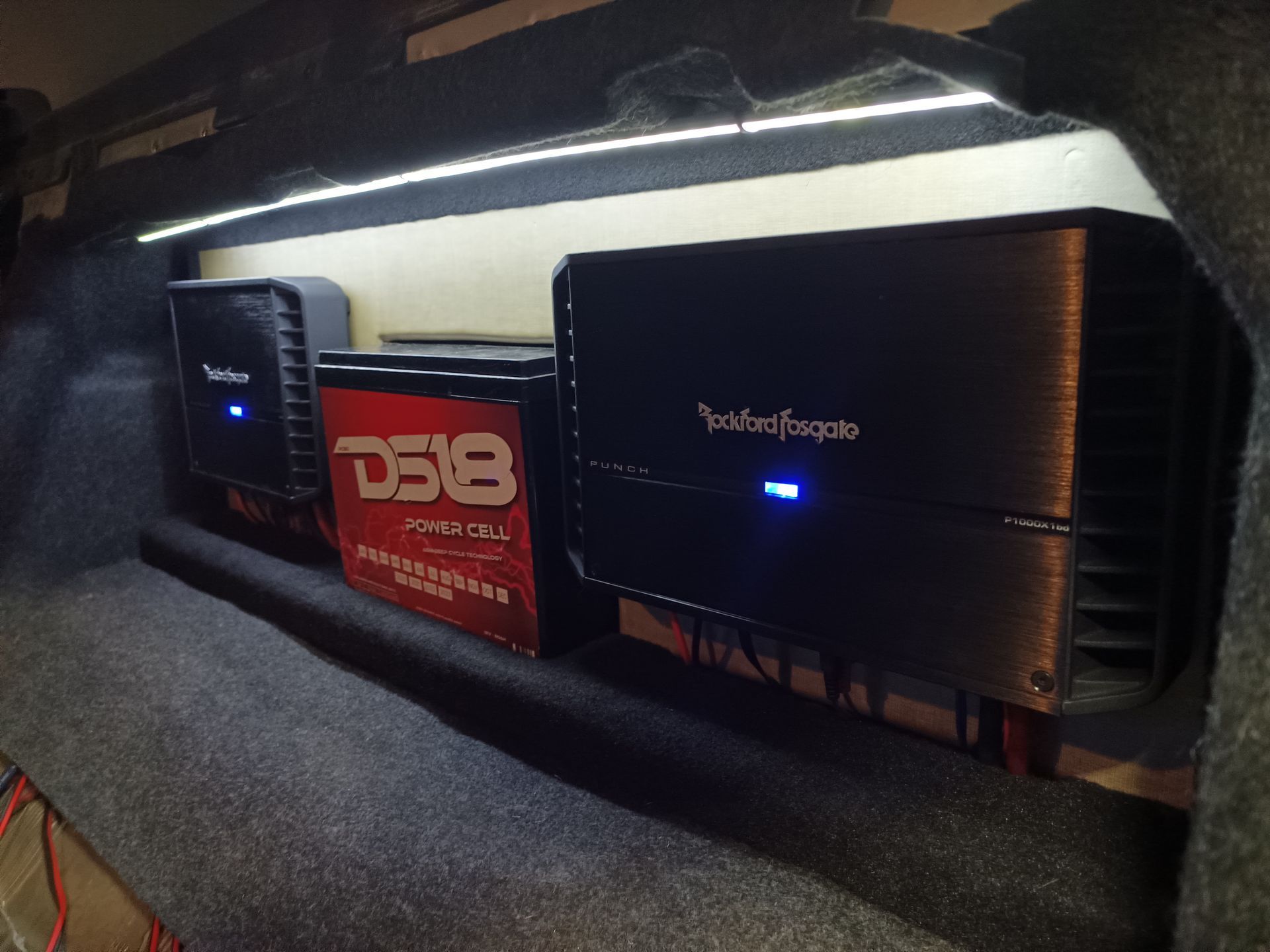 Car audio amplifiers and battery in a vehicle trunk with LED lighting.