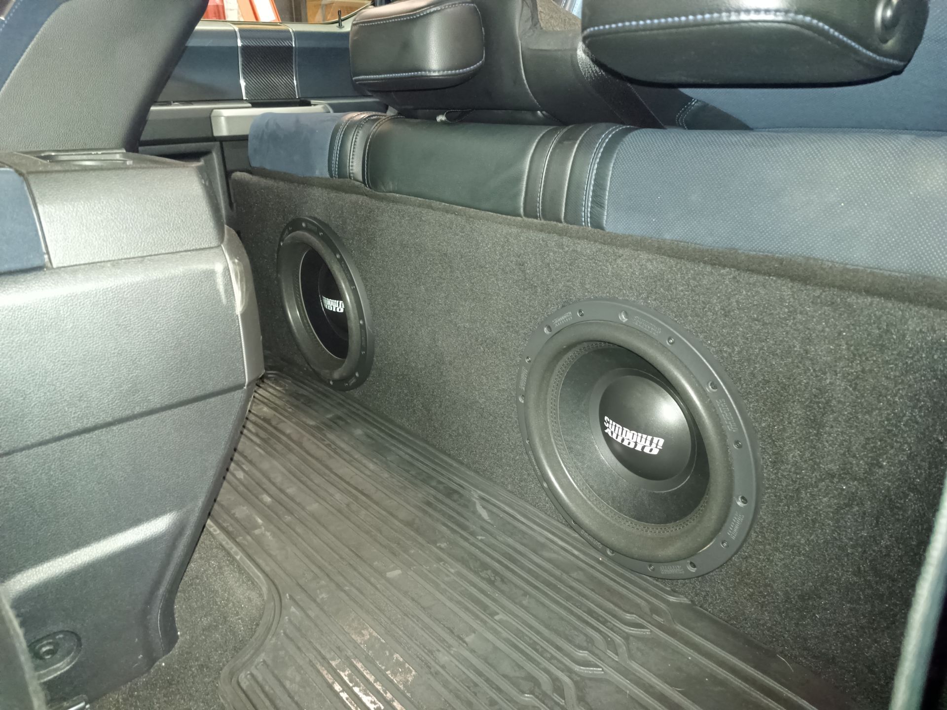 Two black subwoofers built into a black speaker box, installed in a vehicle's back seat.