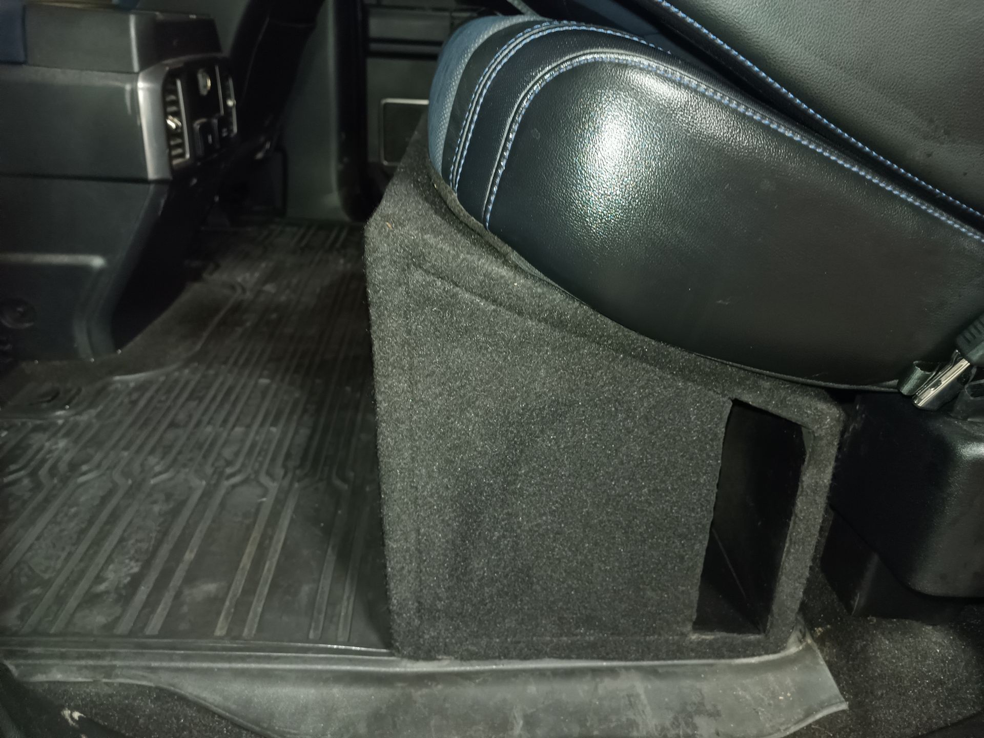 Black subwoofer box in a vehicle, under a black leather seat, near a black floor mat.