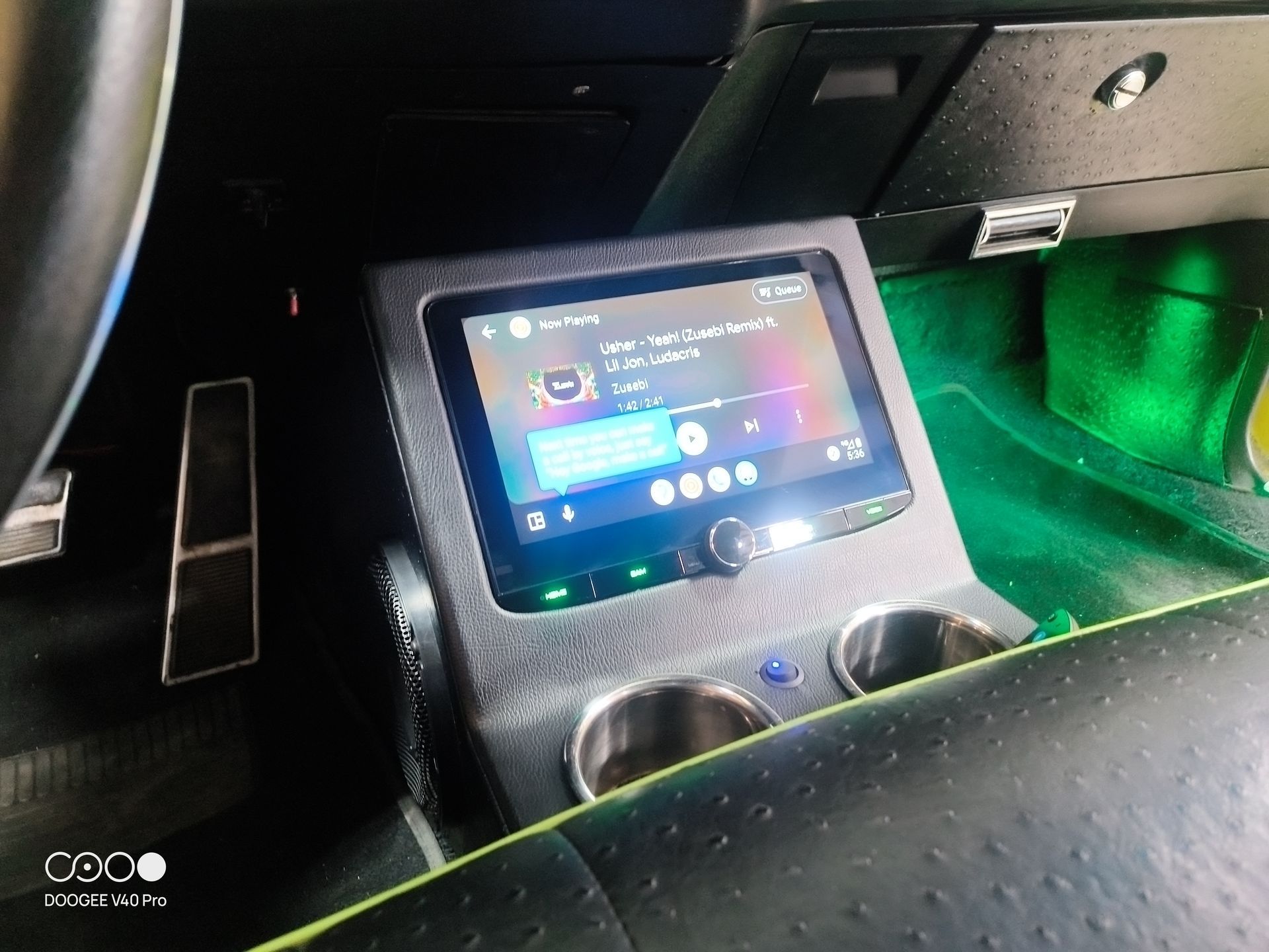 Car's custom console with a screen displaying a music app. Features cup holders and green accent lighting.