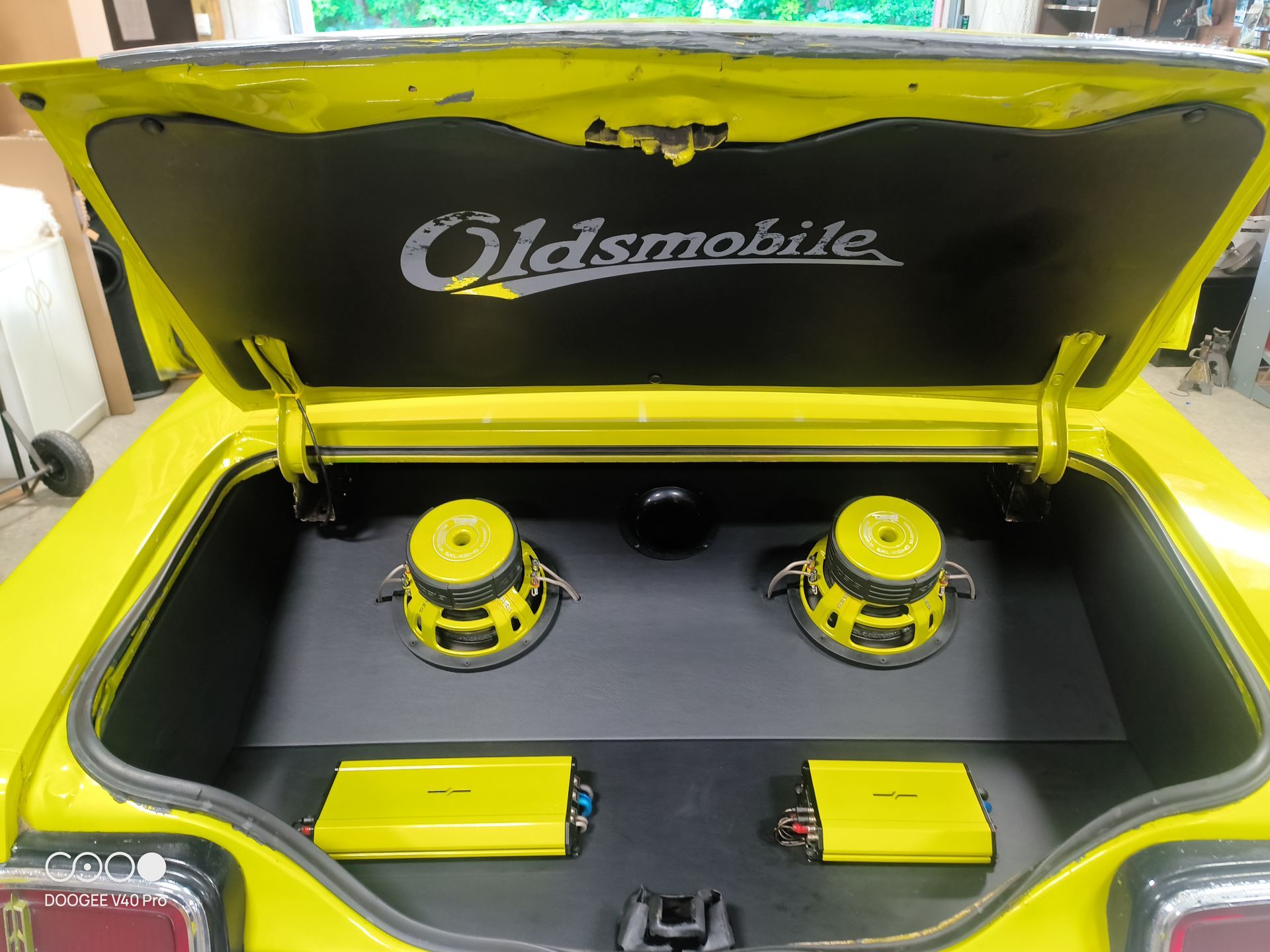Yellow car trunk with Oldsmobile logo, two speakers, and two yellow amplifiers.