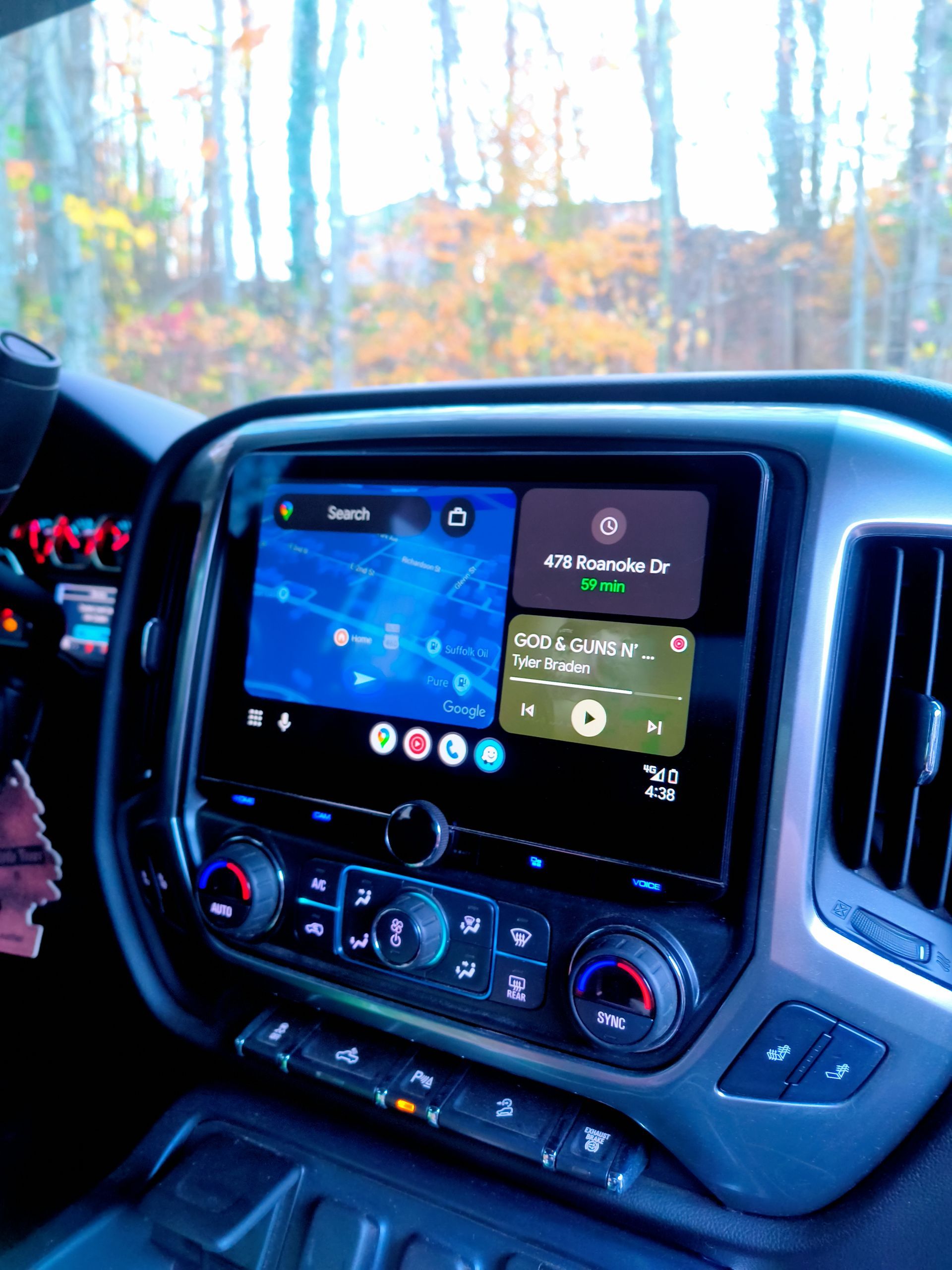 Dashboard with touchscreen display showing map and music controls, inside a vehicle with trees in the background.