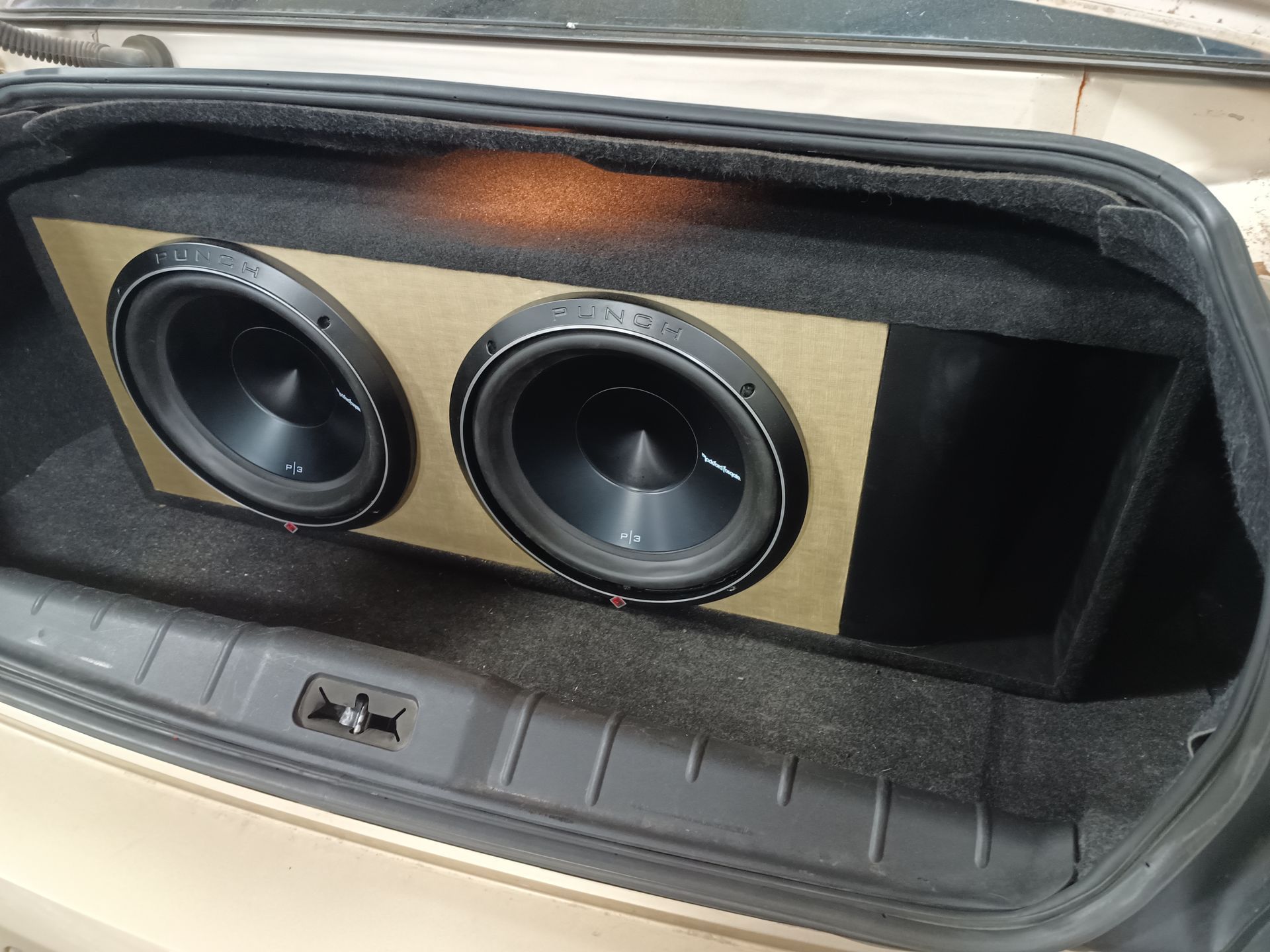 Two black subwoofers mounted in a wooden box inside a car trunk.