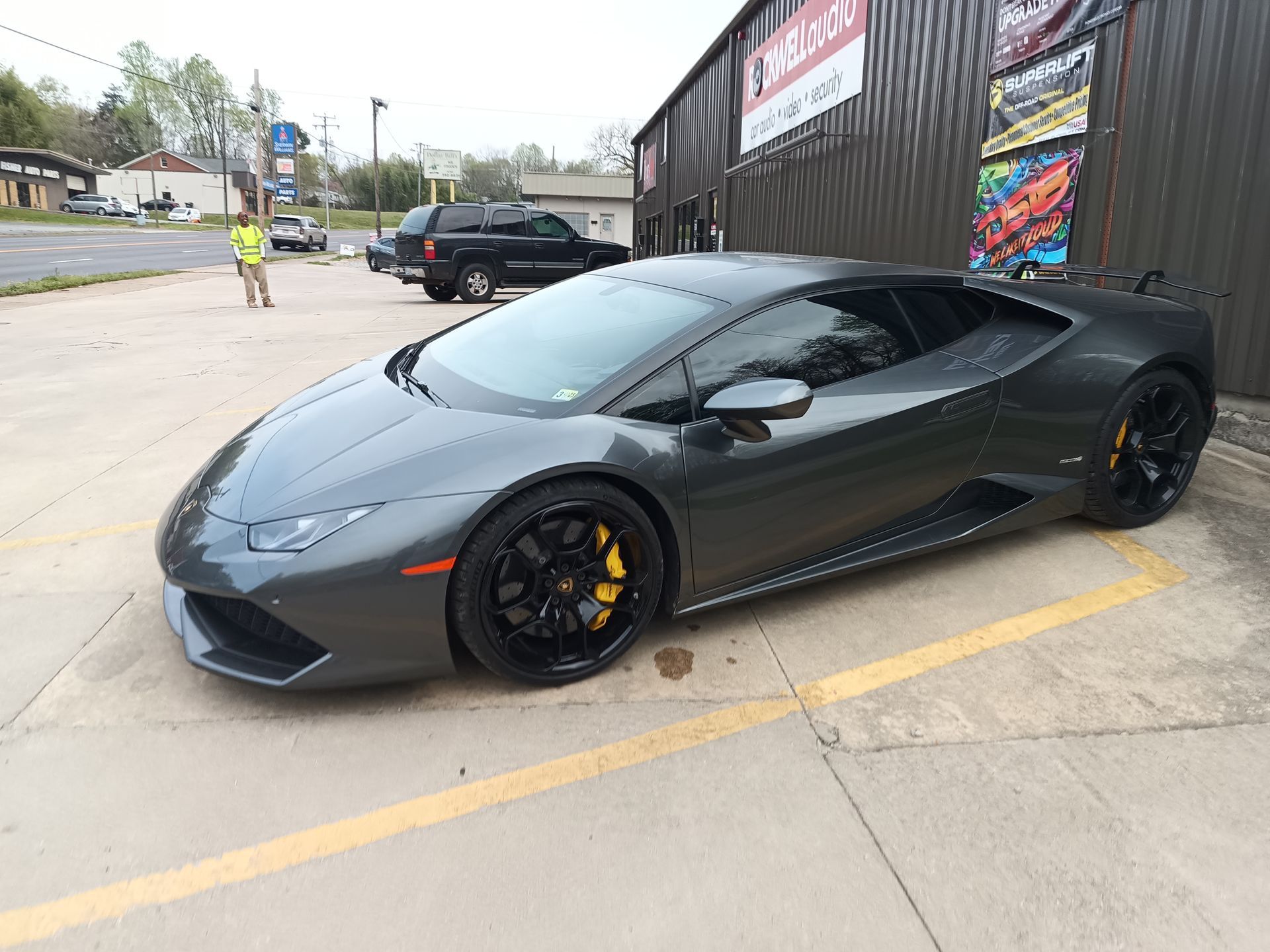 Gray Lamborghini sports car parked in front of a shop; yellow brake calipers, black wheels.