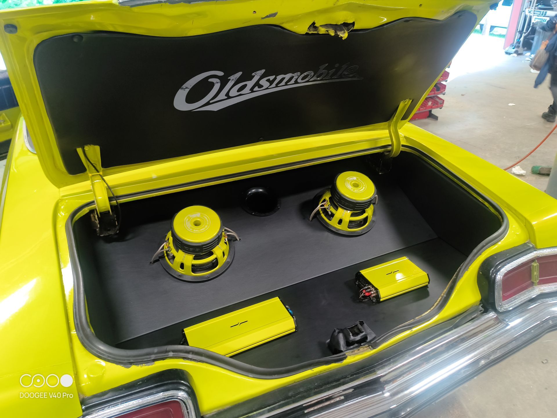 Yellow car trunk with custom stereo, speakers, and amplifiers. Oldsmobile logo.