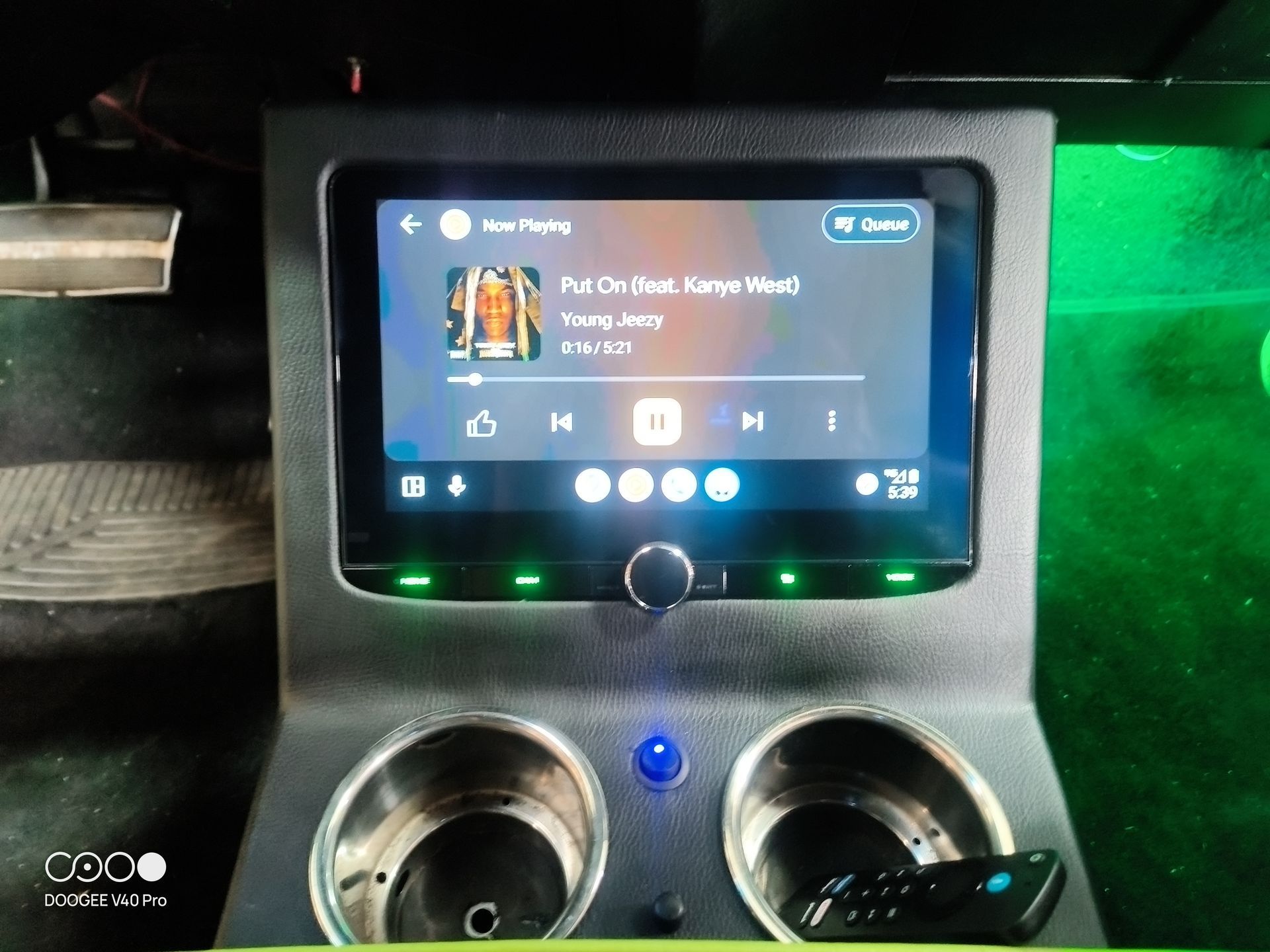 Car's center console with touchscreen display playing music. Cup holders and remote control are visible. Green accent lighting.
