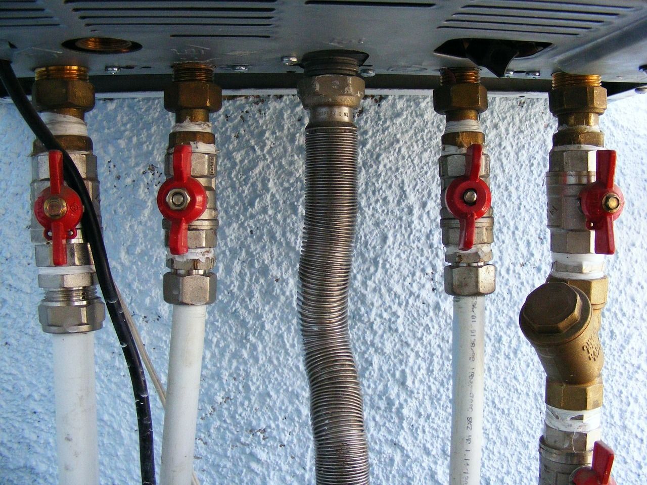 Close-up of plumbing pipes with red valve handles, including a corrugated metal gas line, against a light blue wall.