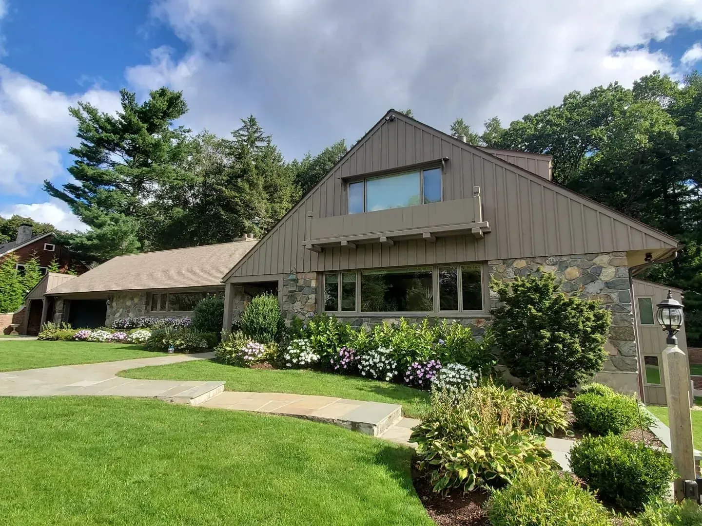 House with stone exterior and tan roof, surrounded by greenery under a cloudy sky.