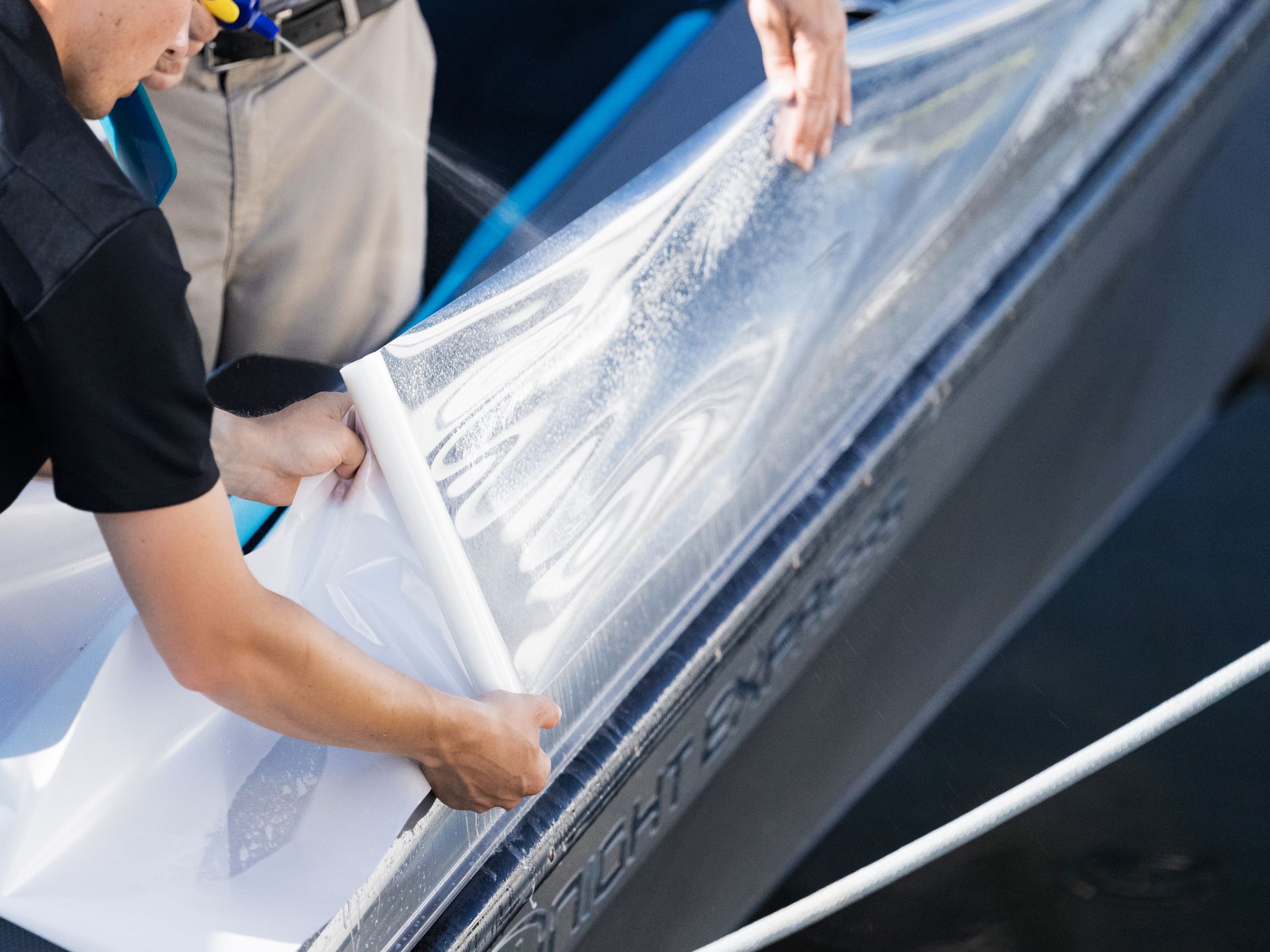 Two people applying clear protective film to a dark boat hull, using water spray.