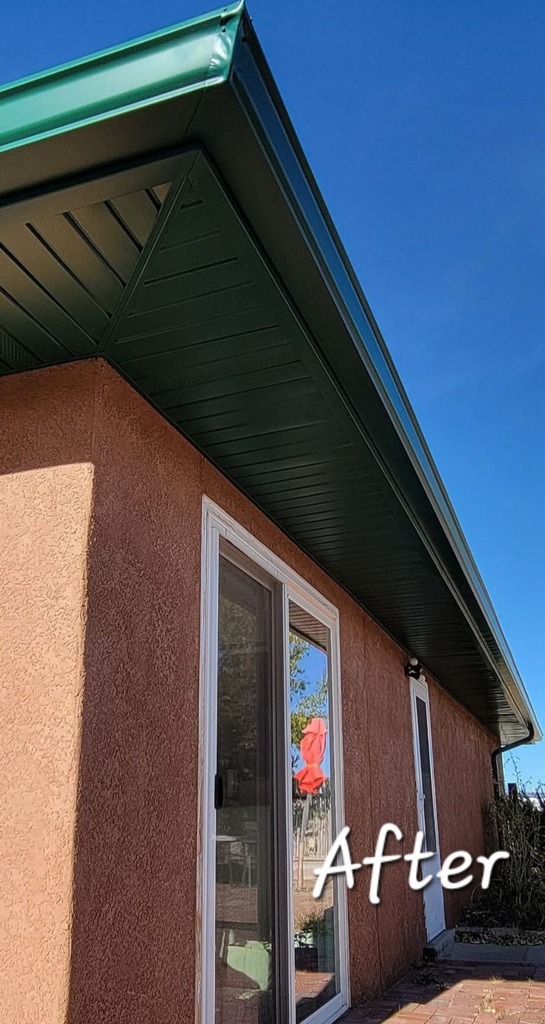 Exterior of a house with brown stucco walls, white-trimmed windows, and a green roof overhang against a blue sky
