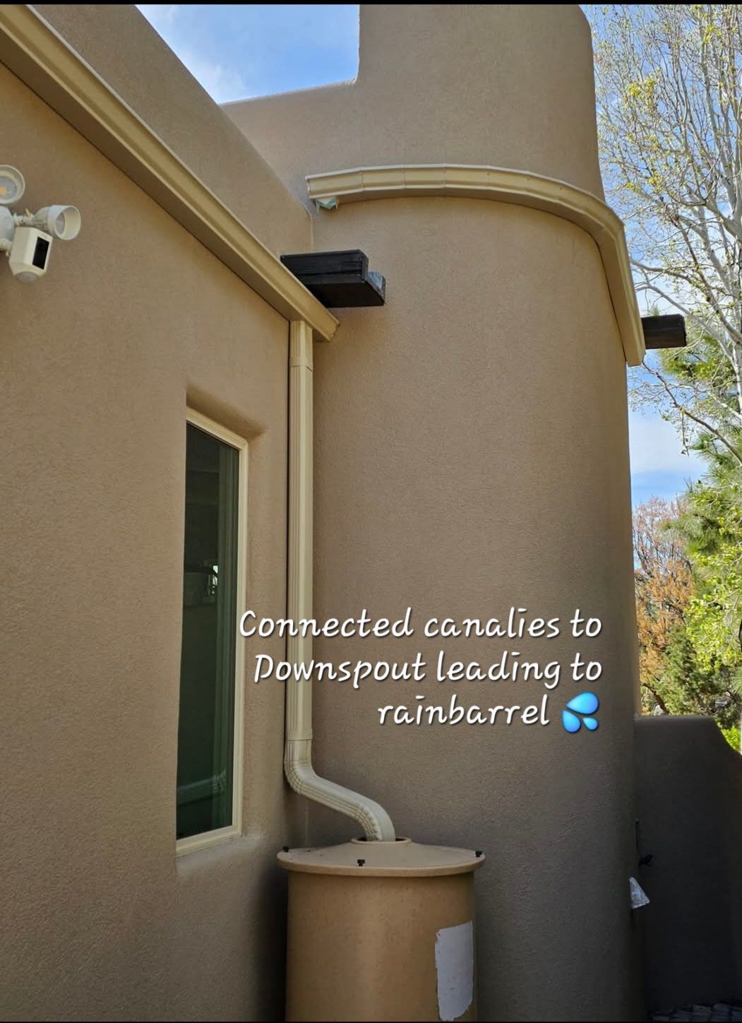 Tan stucco building with a connected downspout leading to a tan rain barrel.