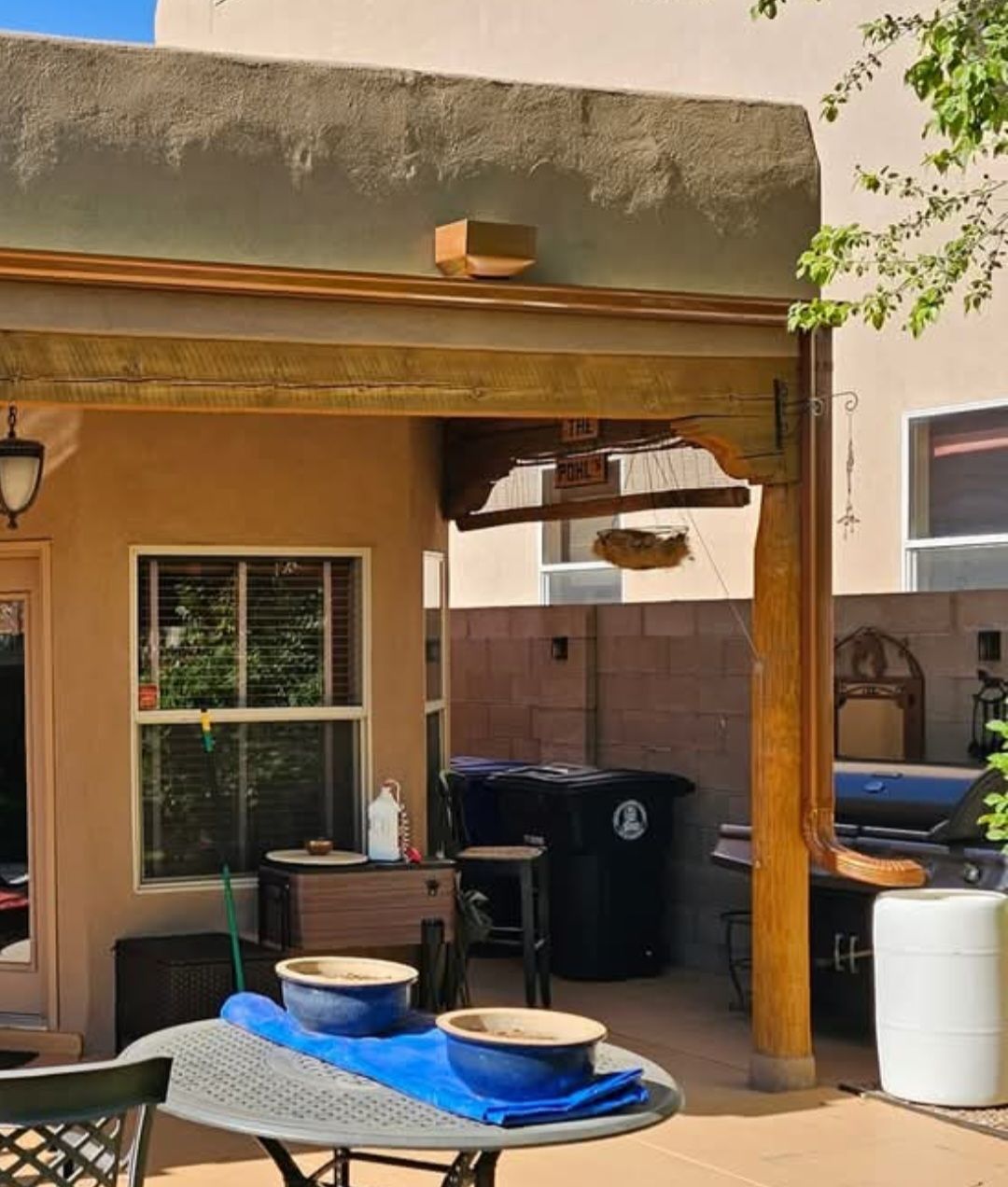 Patio with table and bowls. Brown building, blue bowls, grill, and garbage bins.