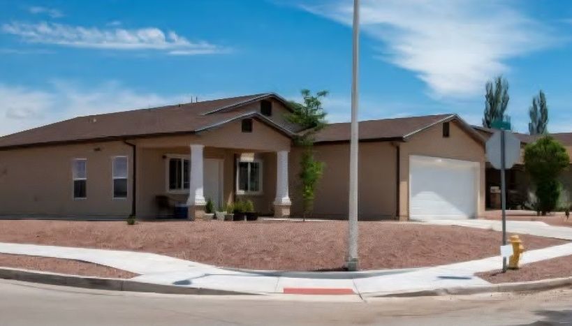 Tan house on a corner lot with a covered porch, garage, and brown roof under a blue sky.