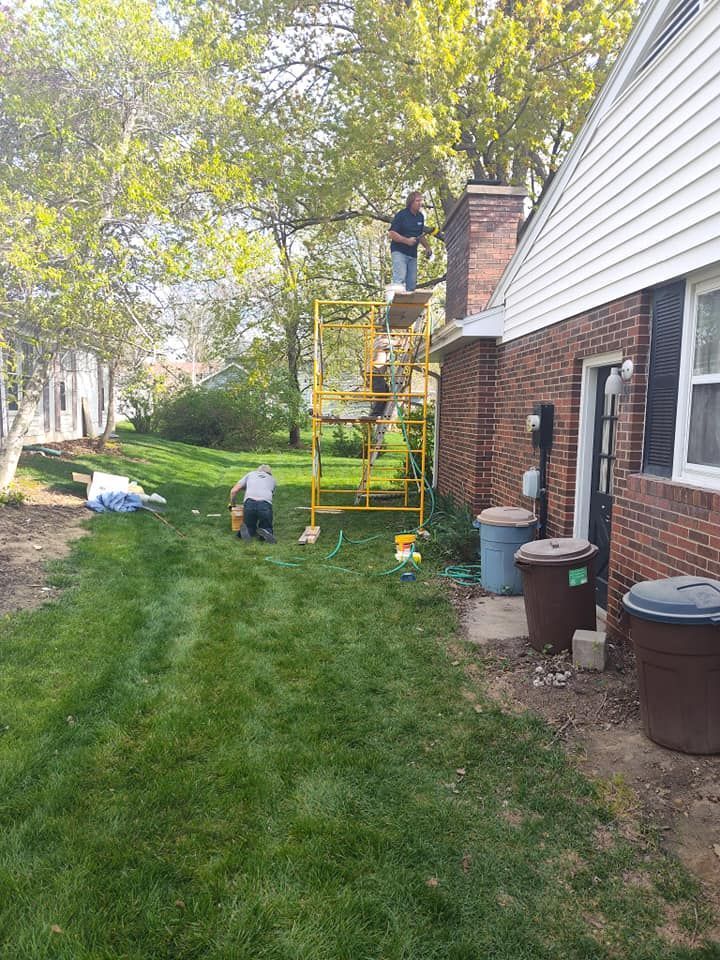 Two workers perform maintenance on a brick chimney using a yellow scaffold in a grassy backyard.