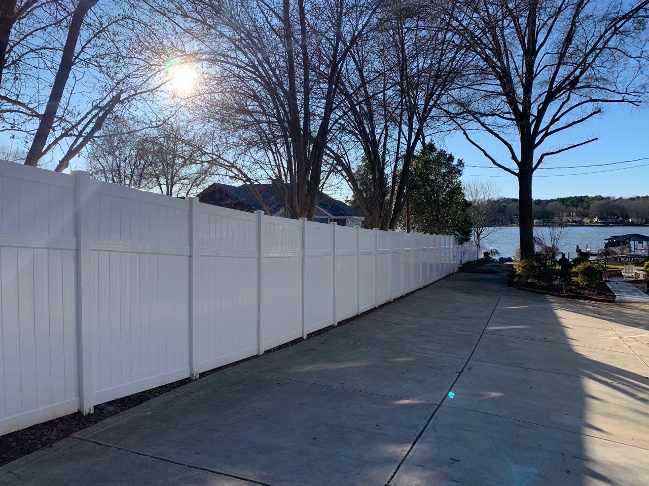 A white fence surrounds a driveway with trees and a lake in the background.