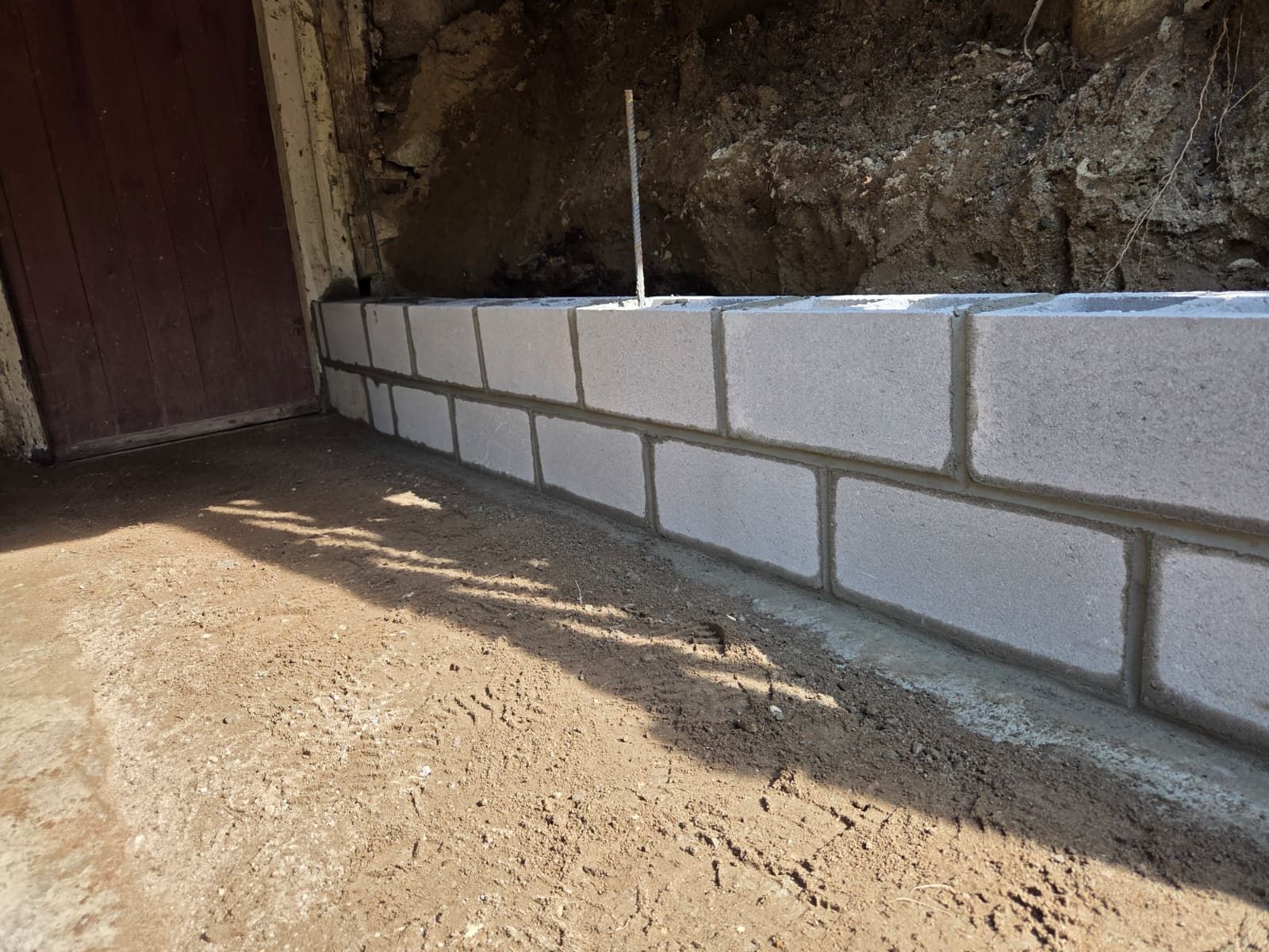Low brick retaining wall beside a dirt path next to a wooden door and rough stone wall.