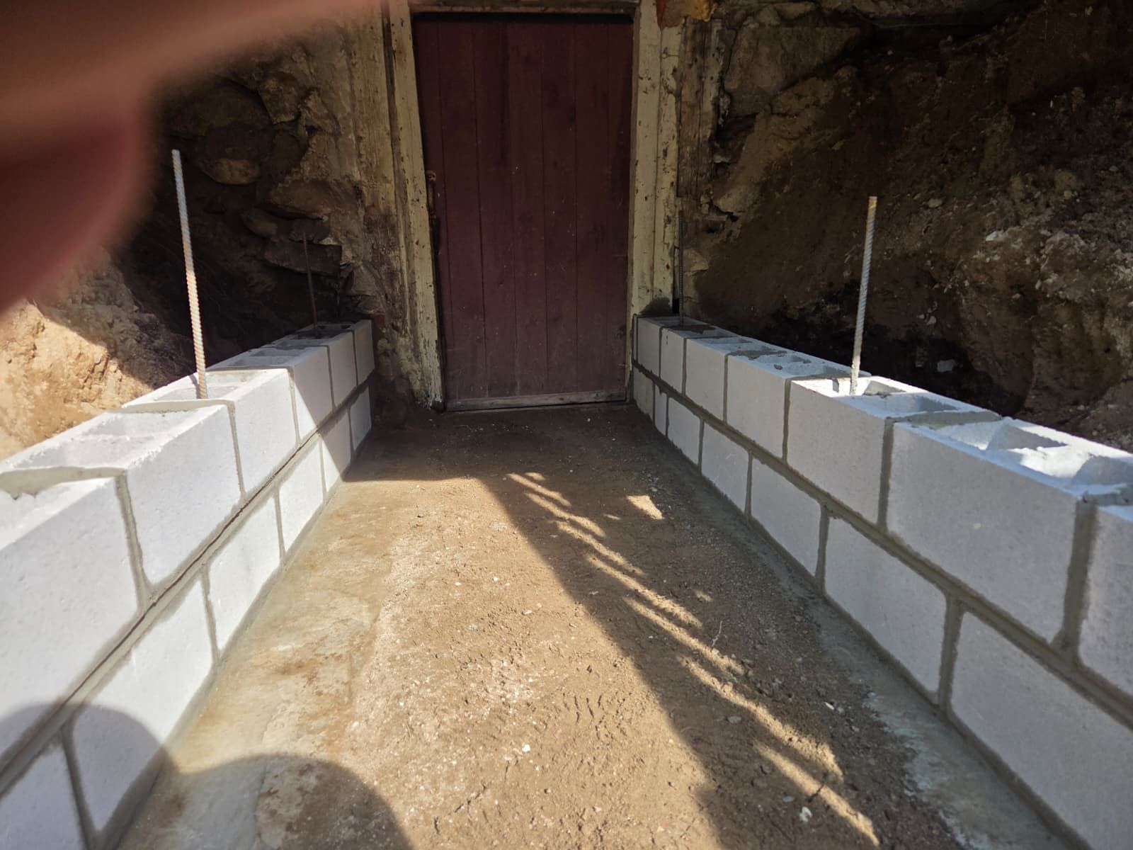 Narrow dirt walkway between cinderblock walls leading to a wooden door in a rocky alcove