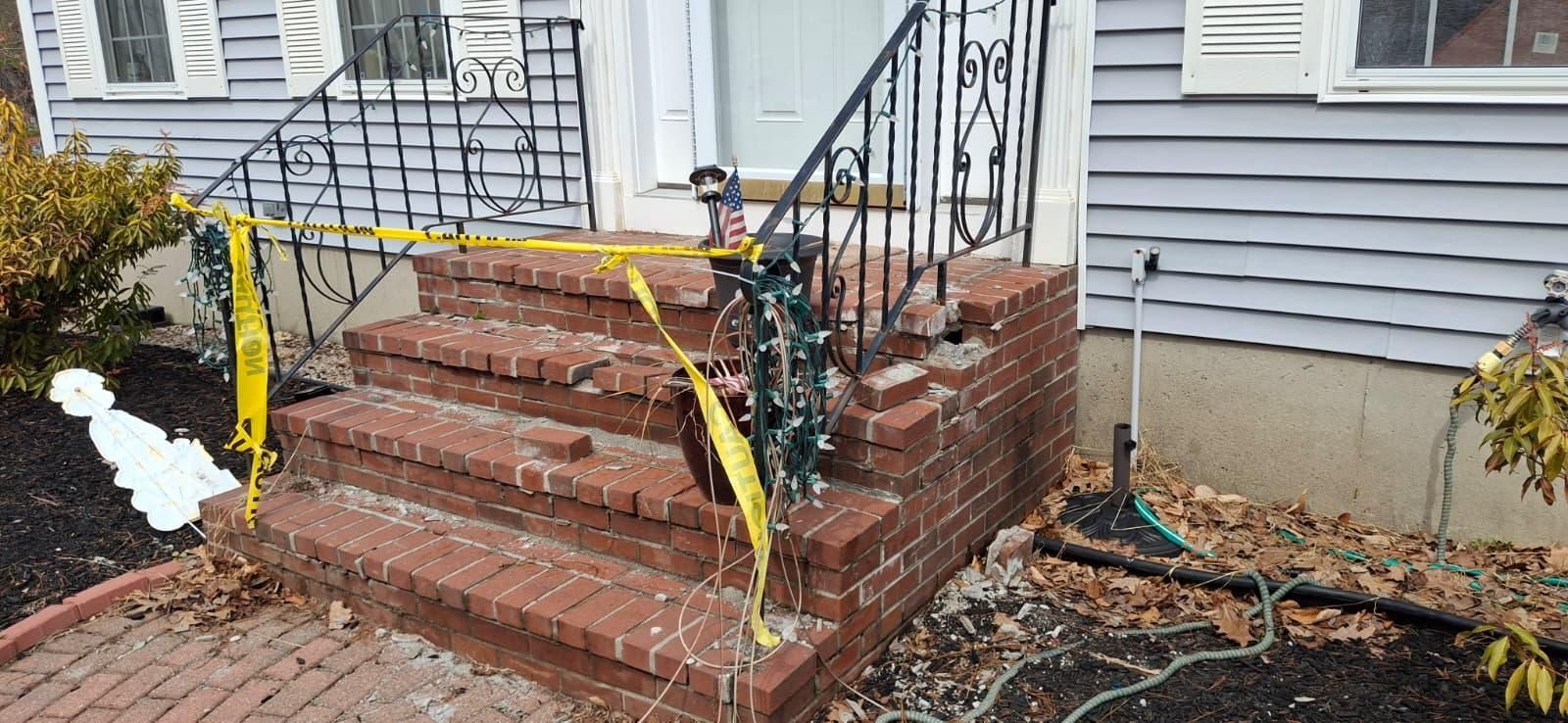 Red brick front steps with black handrail, yellow caution tape, and fallen branches by a house