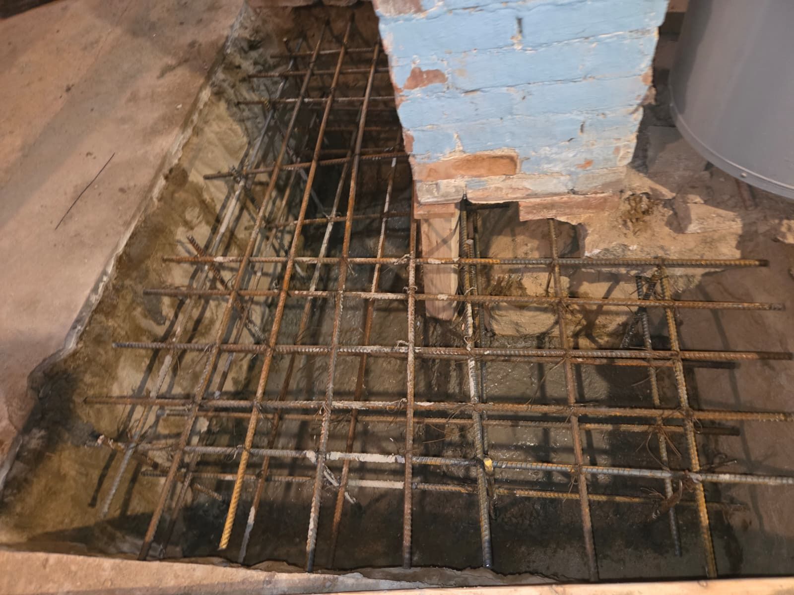 Exposed rebar and broken concrete around a damaged floor opening near a blue wall