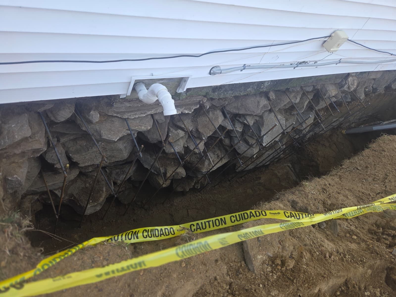 Damaged house foundation with exposed stone wall, yellow caution tape, and dirt excavation work area