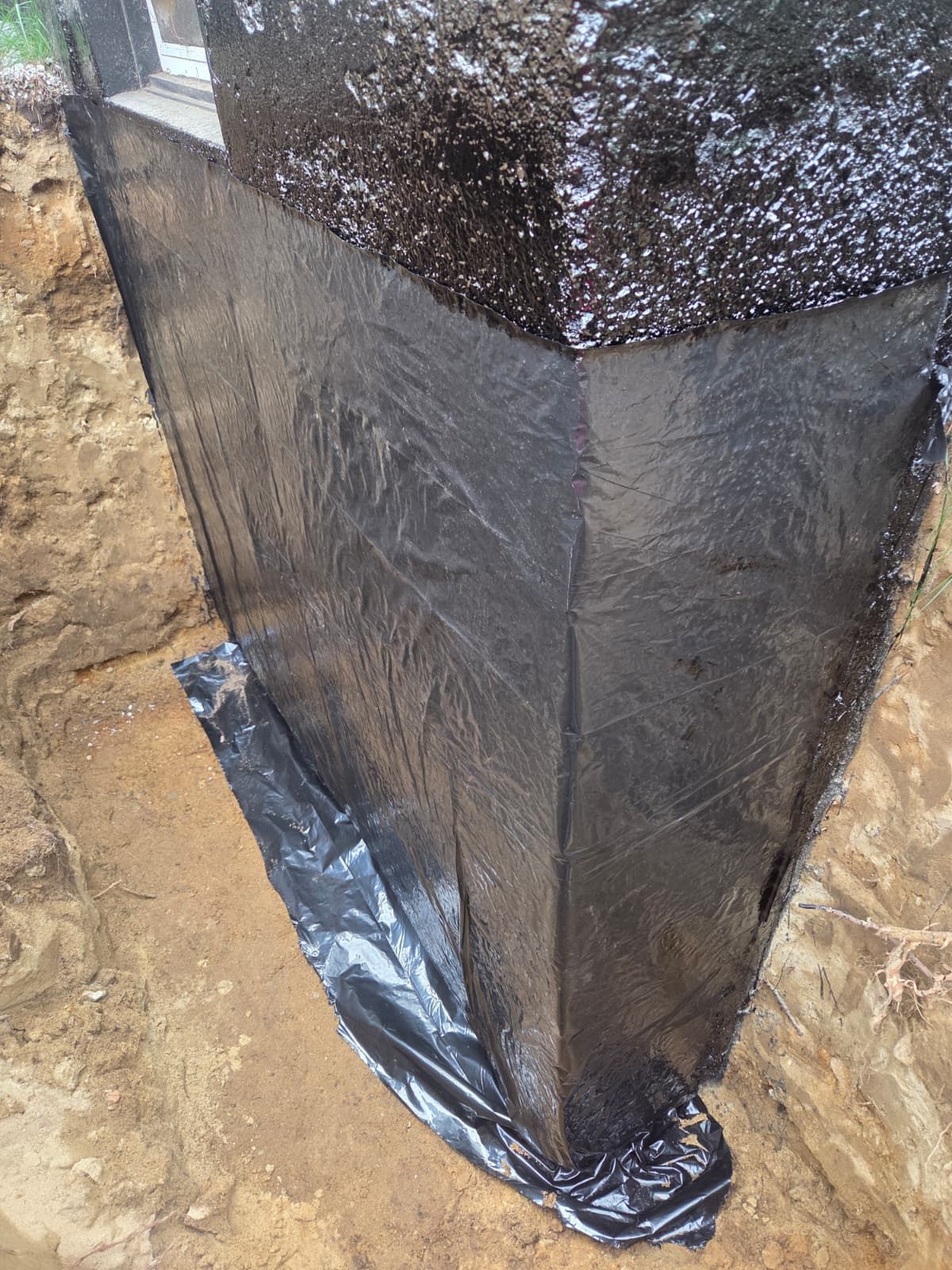 Black waterproofed corner of a building foundation with plastic sheeting at ground level