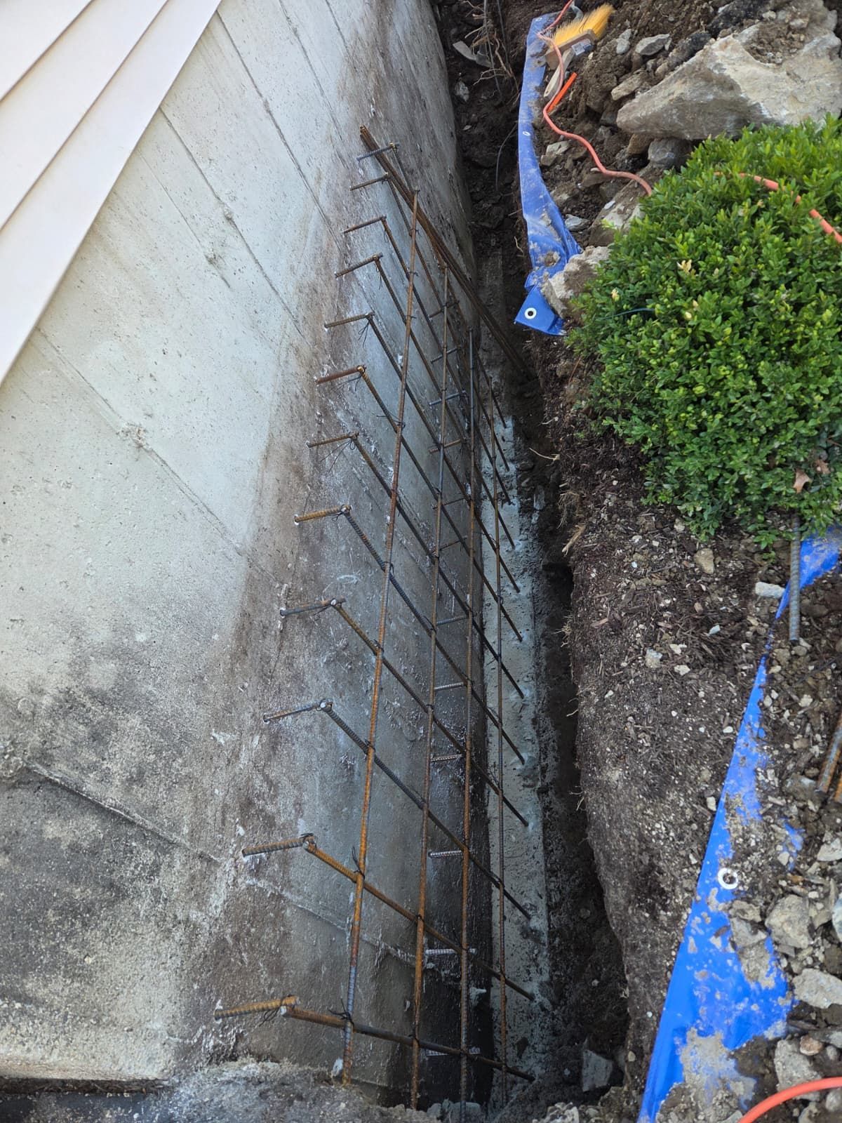 Narrow concrete trench beside a house, lined with rebar, next to dirt, shrubs, and blue tarp.