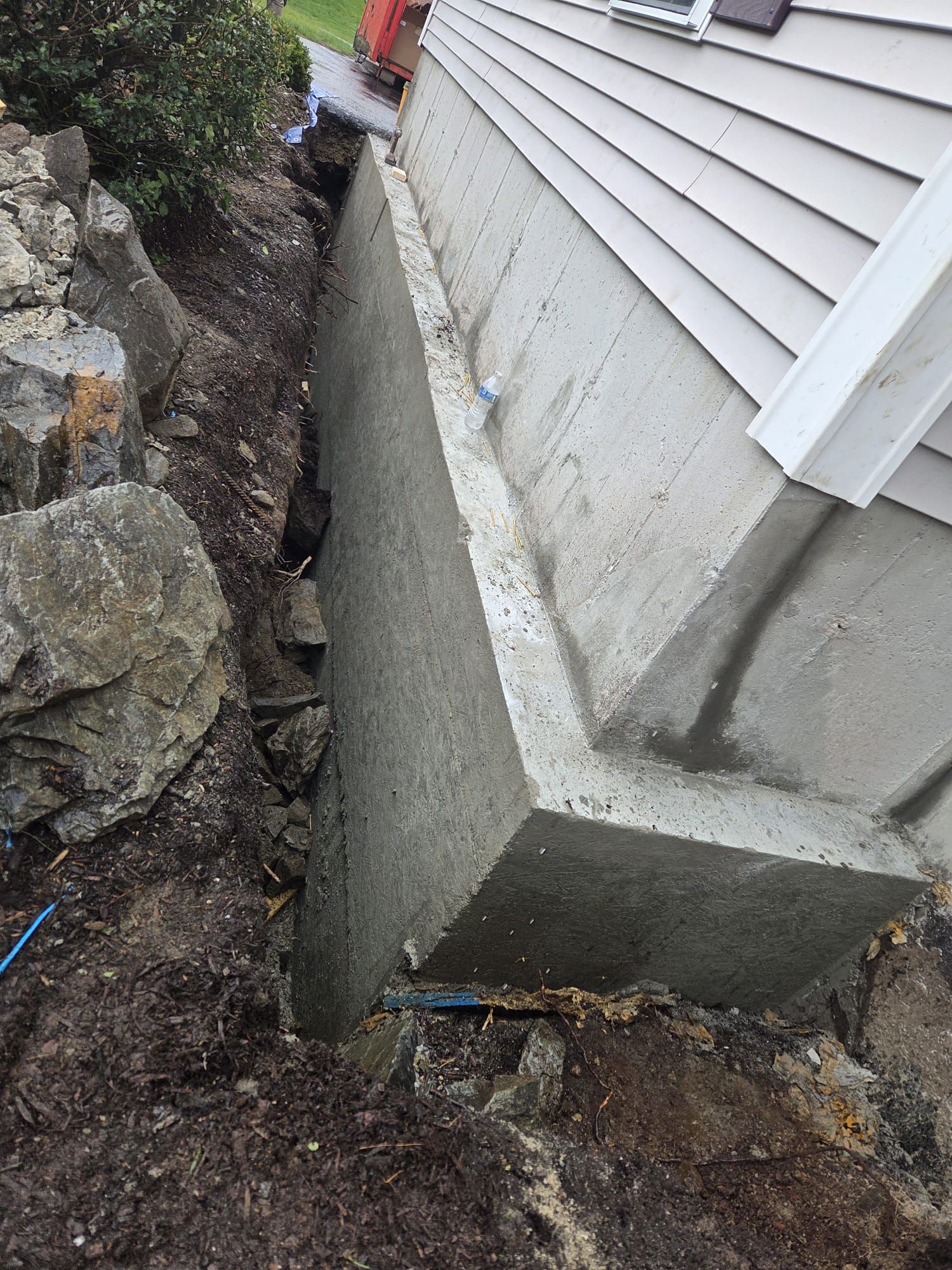 Narrow concrete gap between a house foundation and a rock retaining wall, with dirt and debris below.