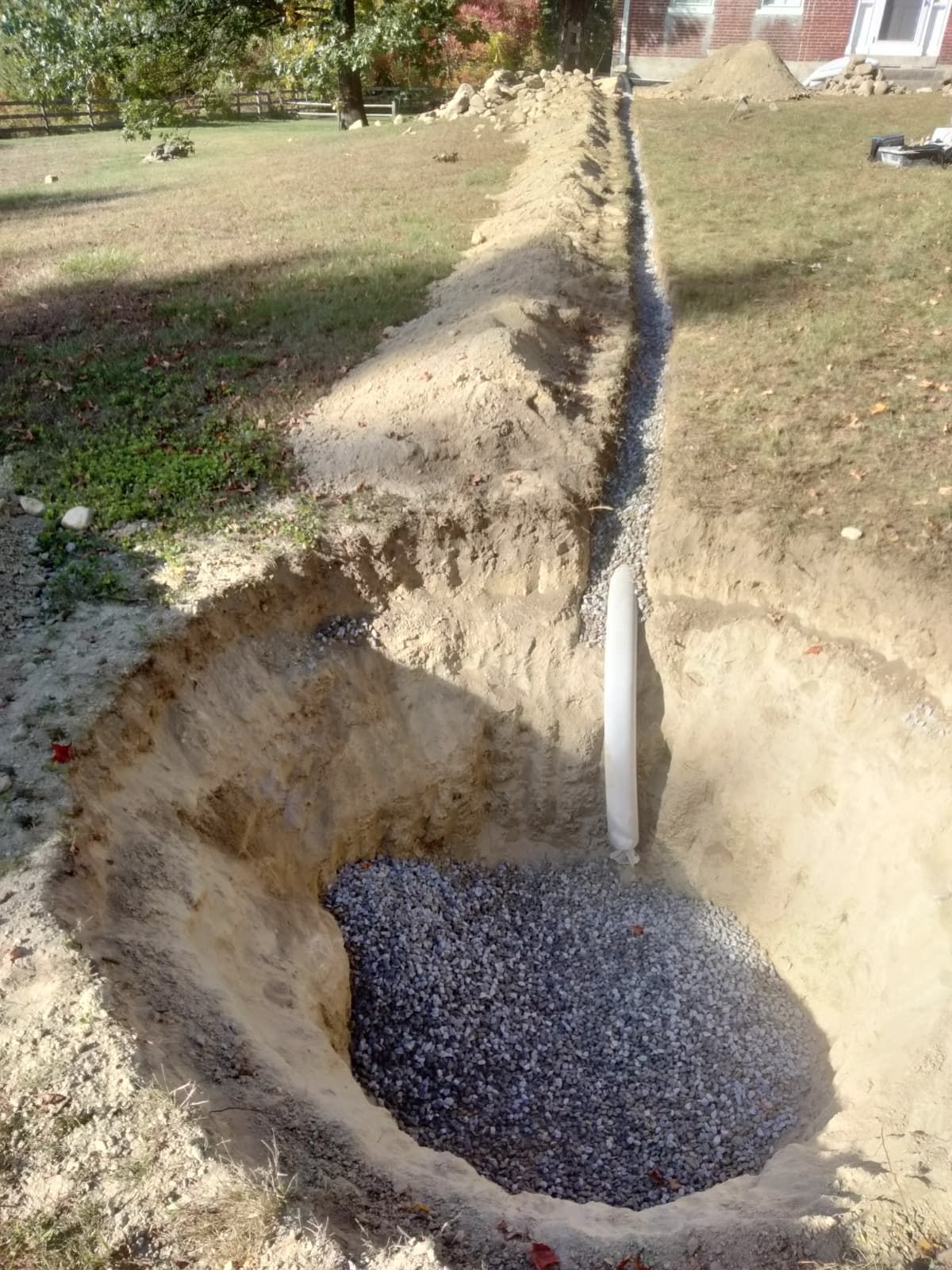 Trench with a white pipe pouring gravel into a large excavated hole in a yard