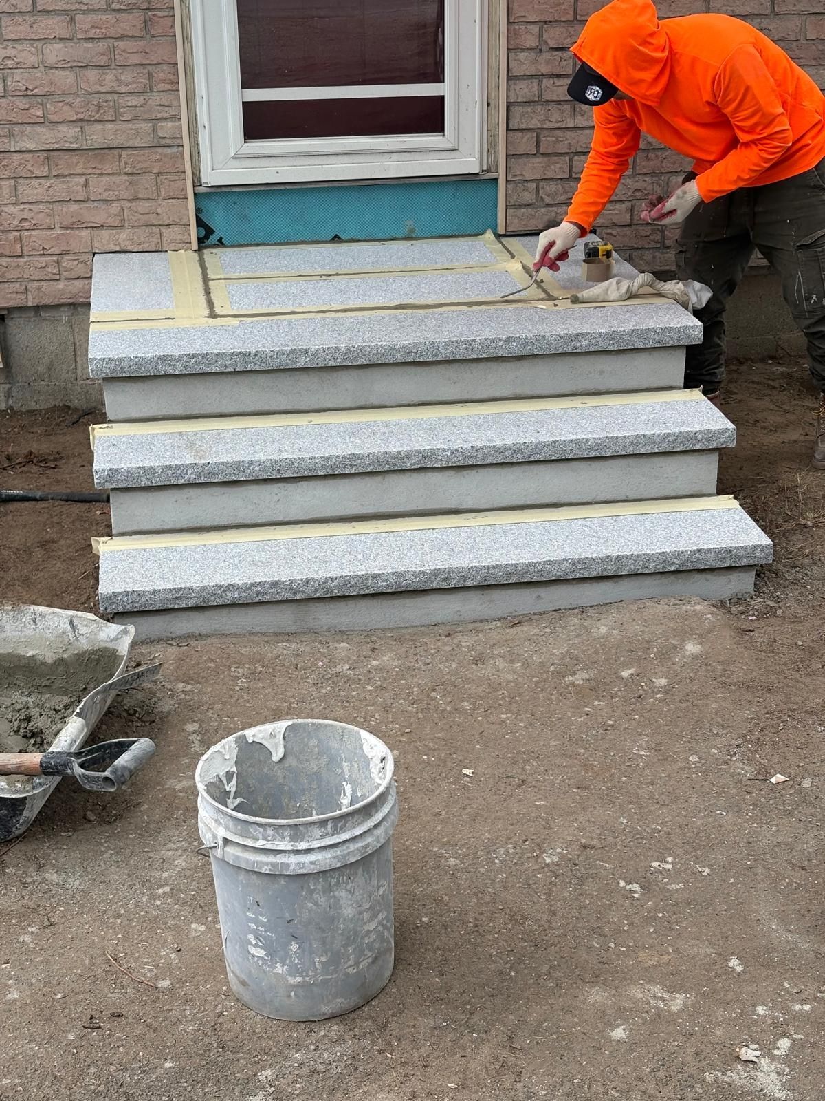Worker in orange hoodie installing stone steps on a house stoop with a bucket in front.