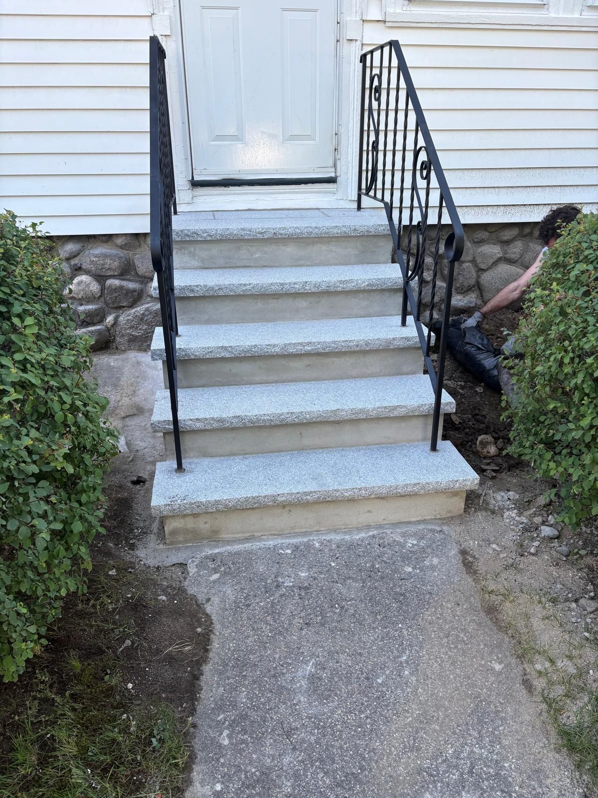 Front steps with black railings leading to a white house door, bordered by shrubs and stone walls