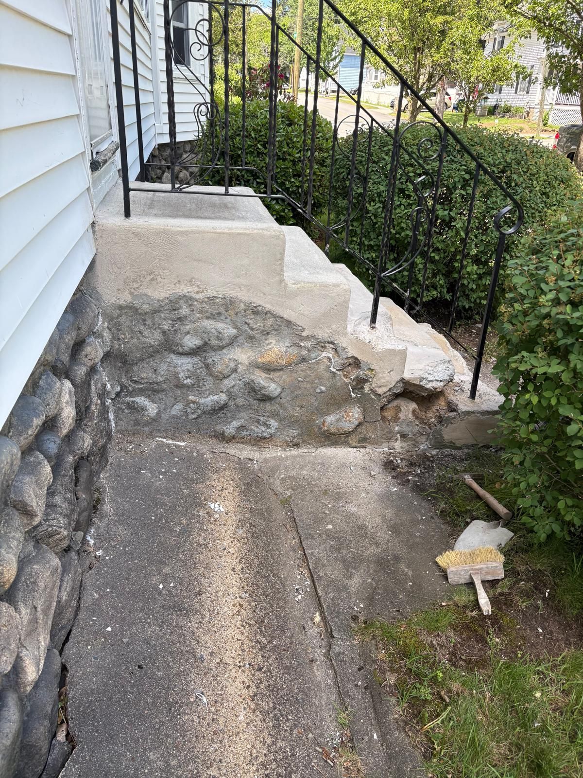 Damaged front steps with crumbling concrete, stone foundation, and black iron railing beside a house