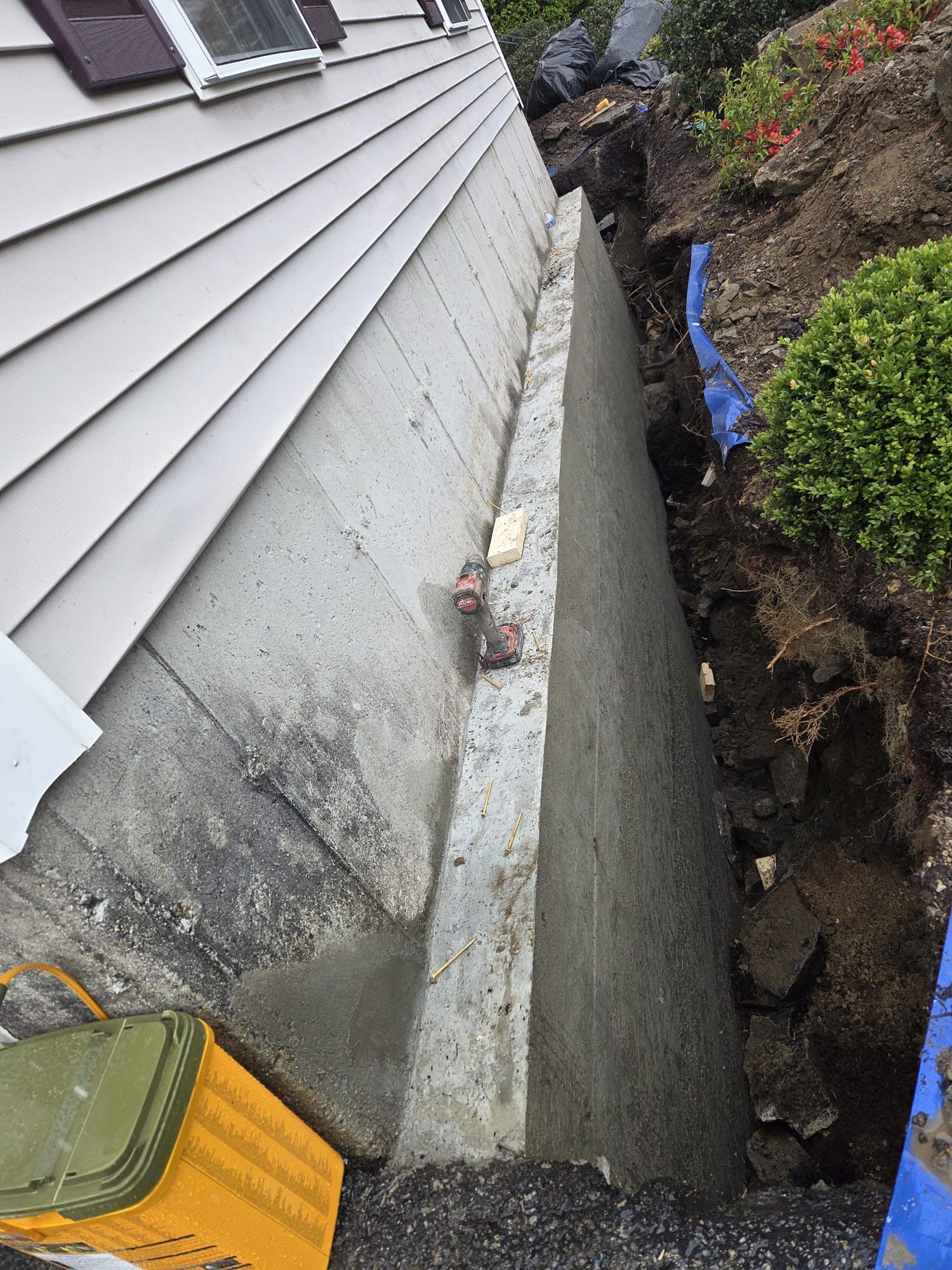 House foundation beside a narrow trench with exposed soil and a blue pipe, next to gray siding.