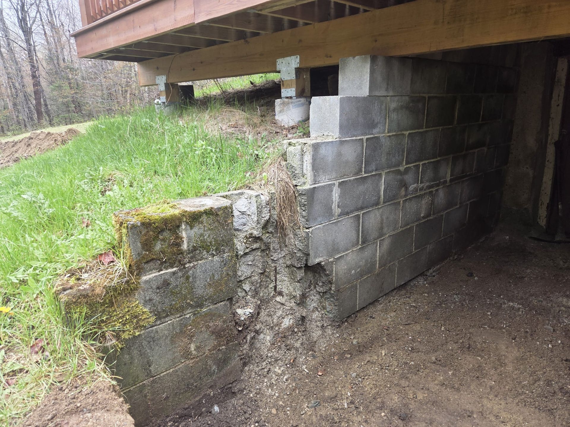 Moss-covered cinder block foundation under a raised deck, with a damaged corner beside grassy ground.