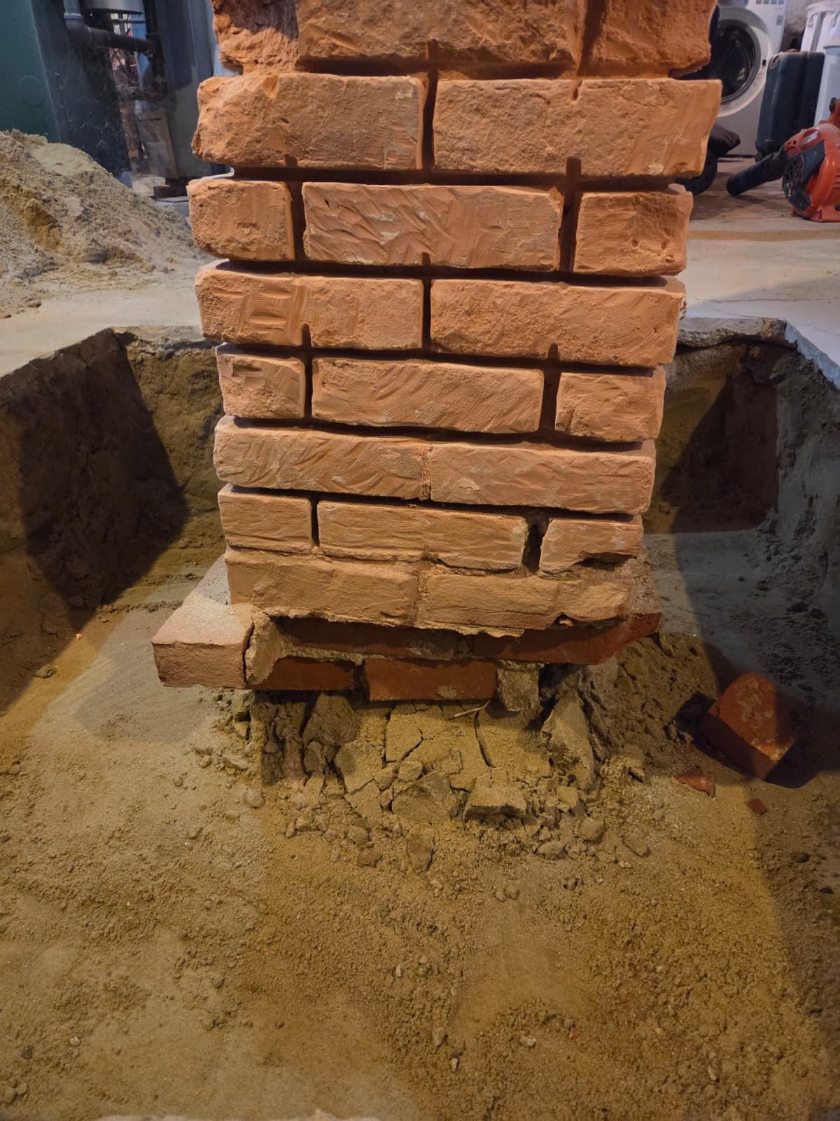 Stacked red bricks on muddy ground, with a damaged base at the bottom of the wall.