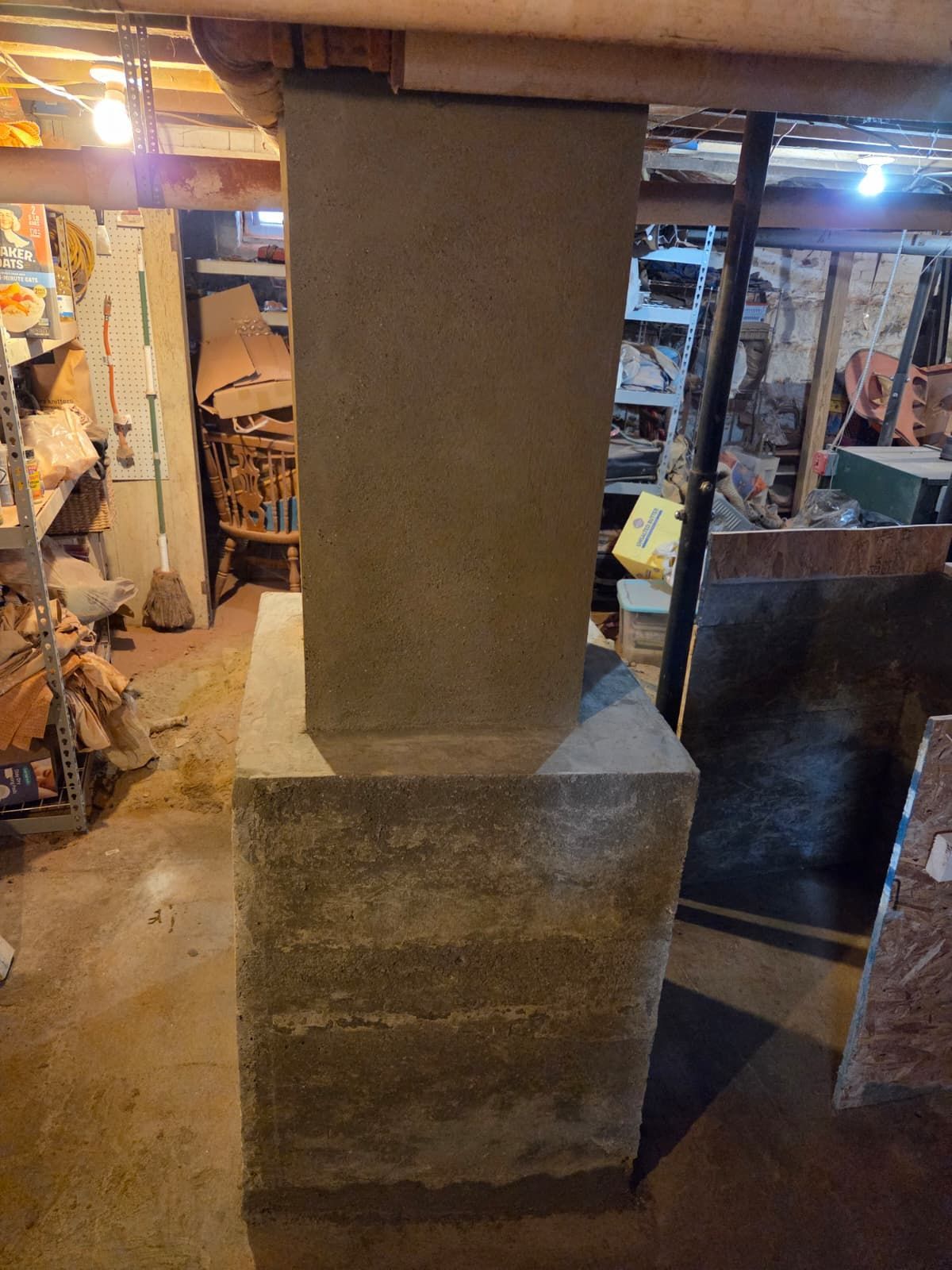 Concrete support pillar in a basement with storage shelves and items in the background