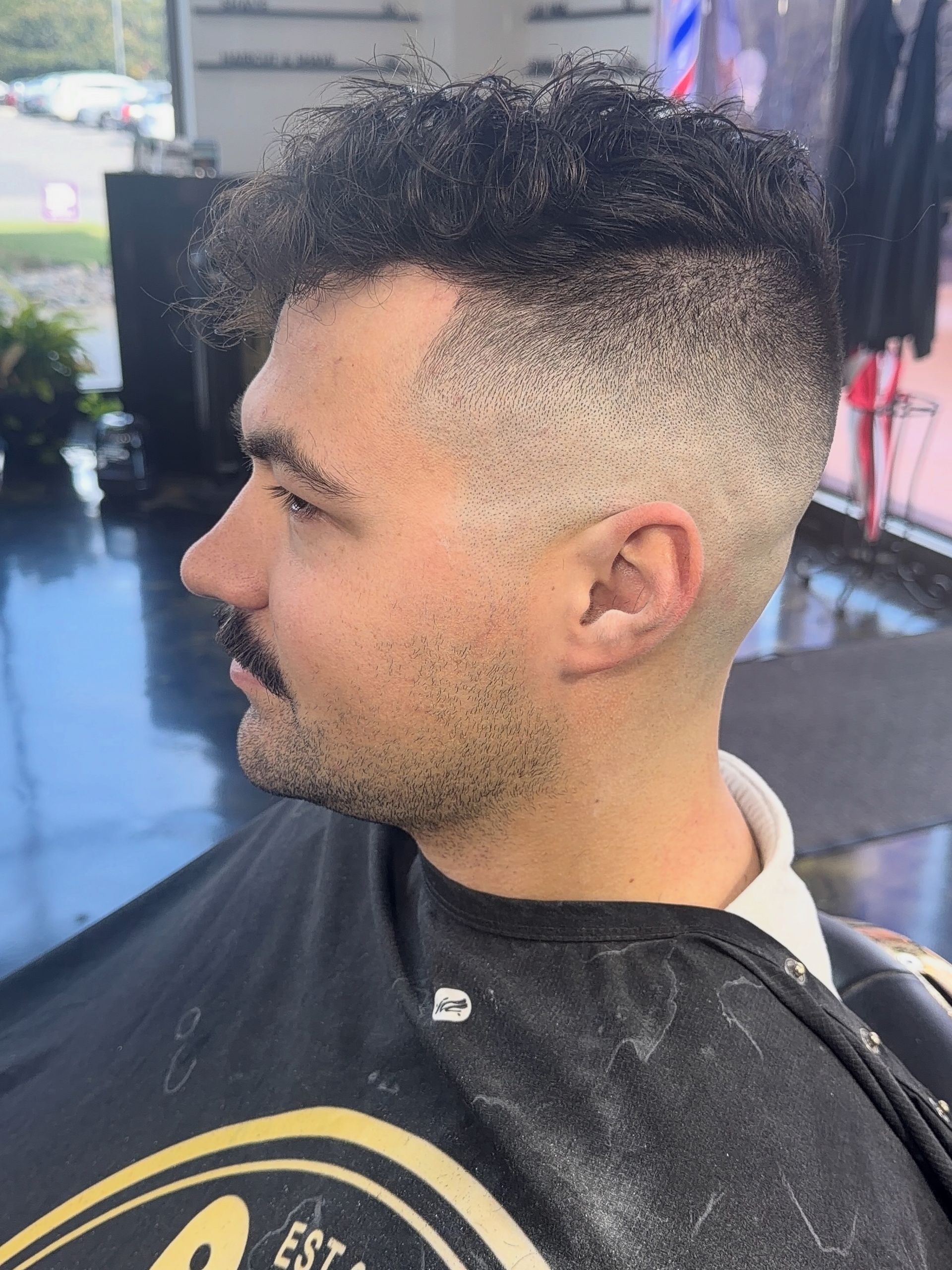 Side-profile portrait of a man with a curly fade haircut, seated in a barber chair wearing a black cape.