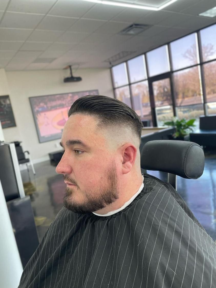Man seated in a barber chair in a bright salon, wearing a dark striped cape, side profile visible.