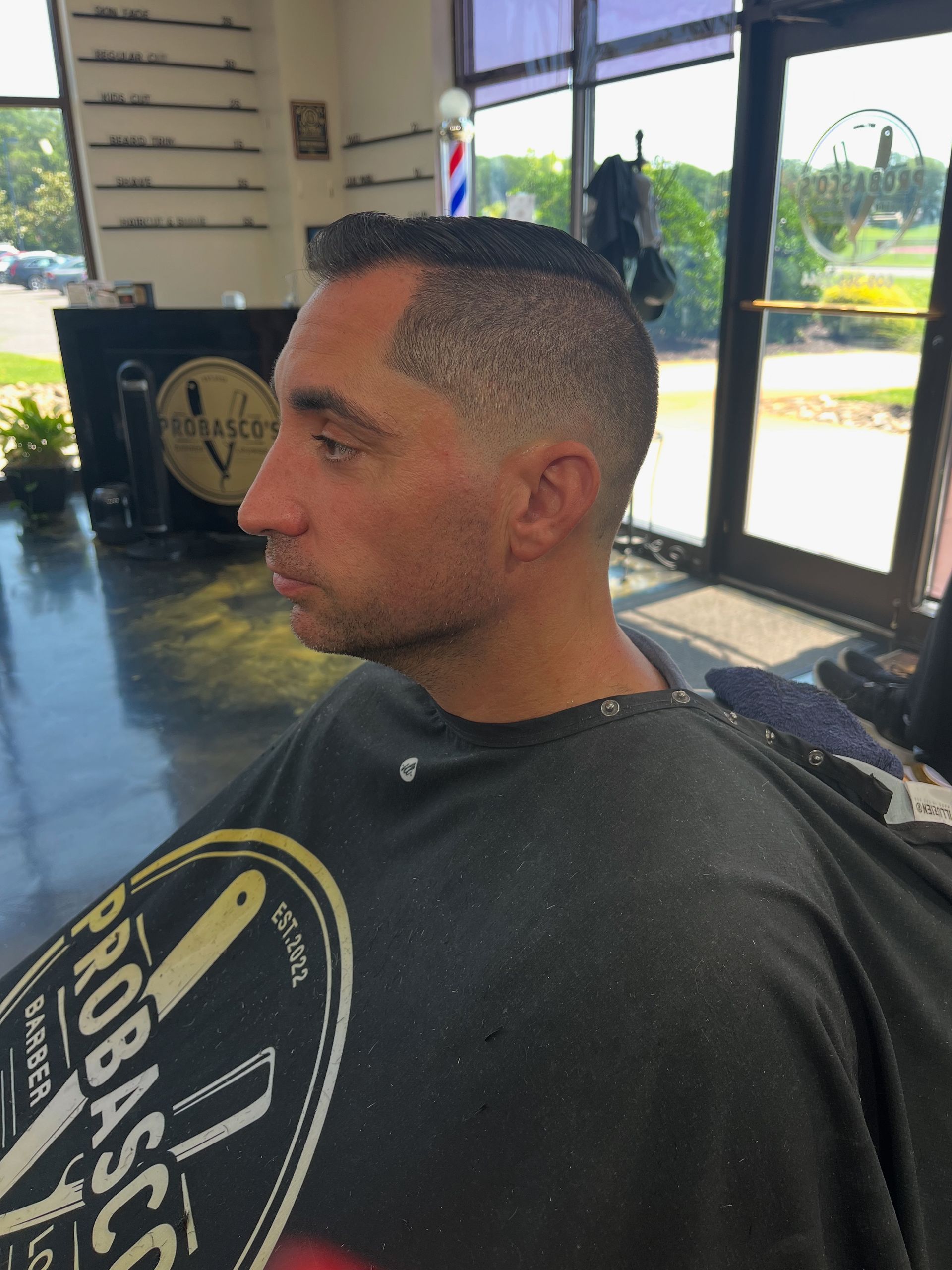 Man with a close fade haircut sitting in a barber chair, wearing a black cape in a bright barbershop.