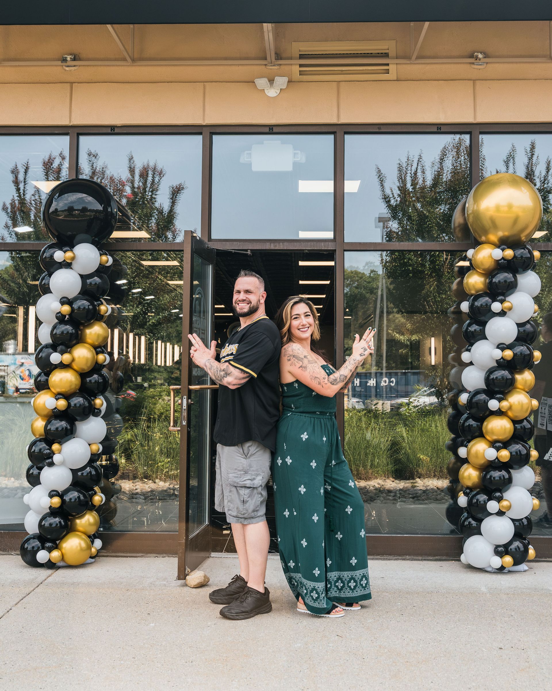 Couple posing outside a storefront between black, white, and gold balloon columns, smiling at the camera