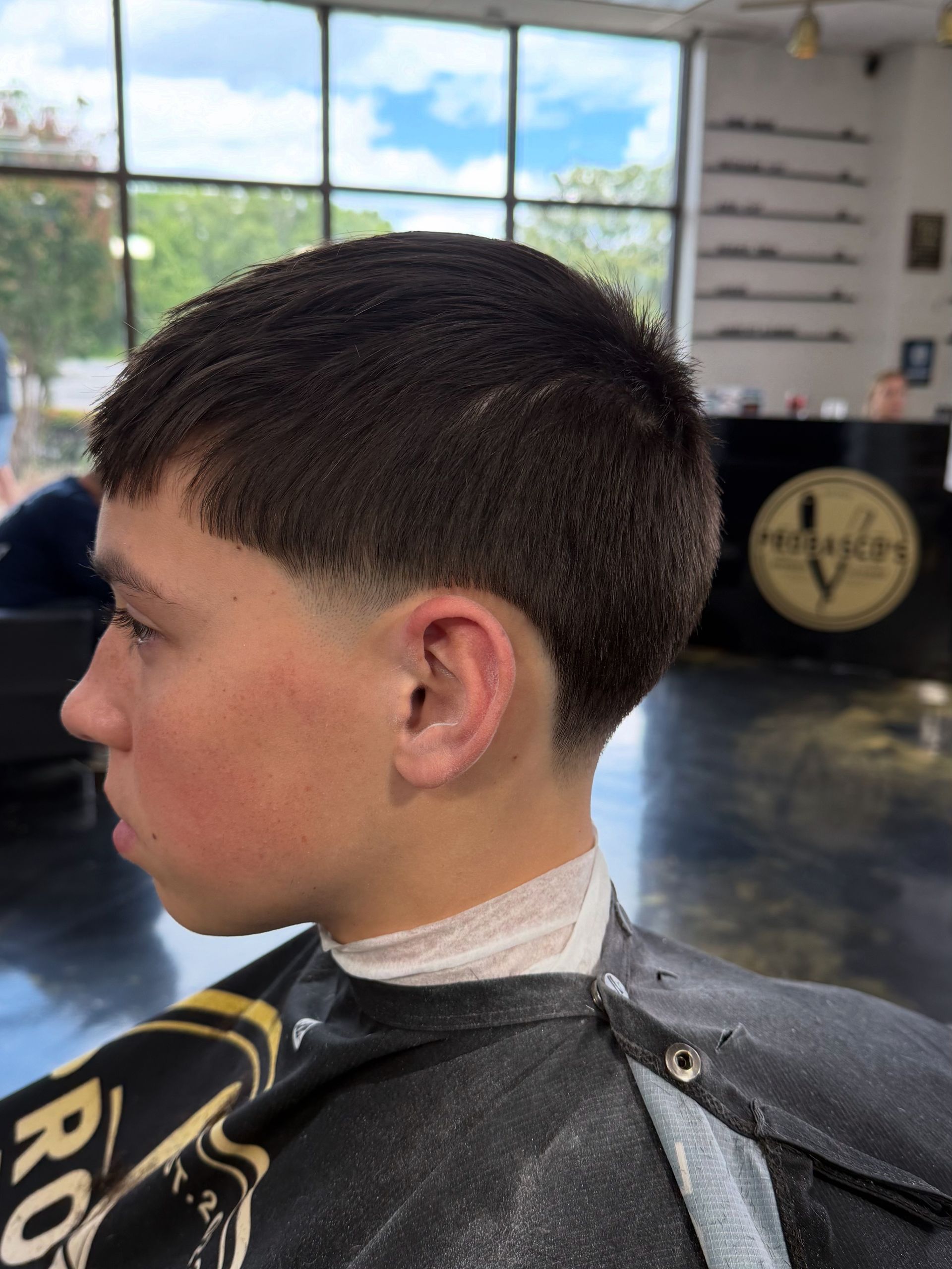 Side profile of a person with a short haircut in a barber chair, wearing a black cape in a bright salon.