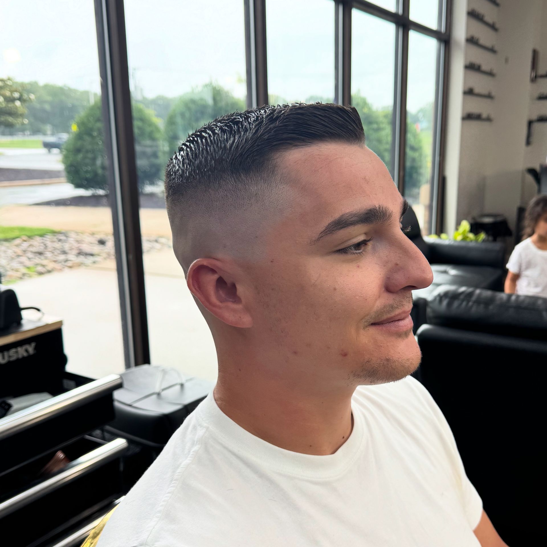 Man with a short fade haircut in a white T-shirt, seated indoors by large windows in a salon.