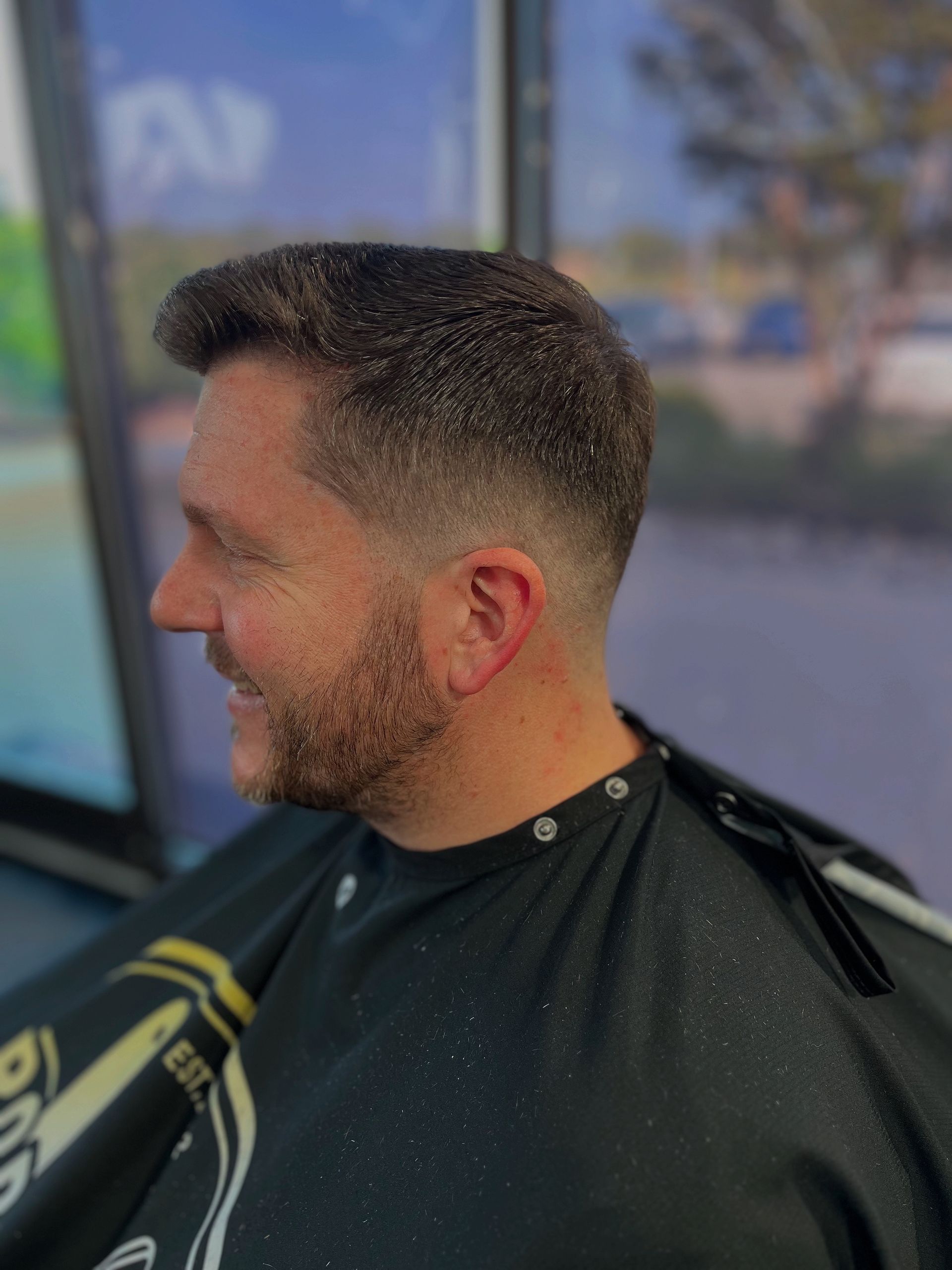 Man with a short fade haircut and beard wearing a black barber cape, smiling in a barbershop chair