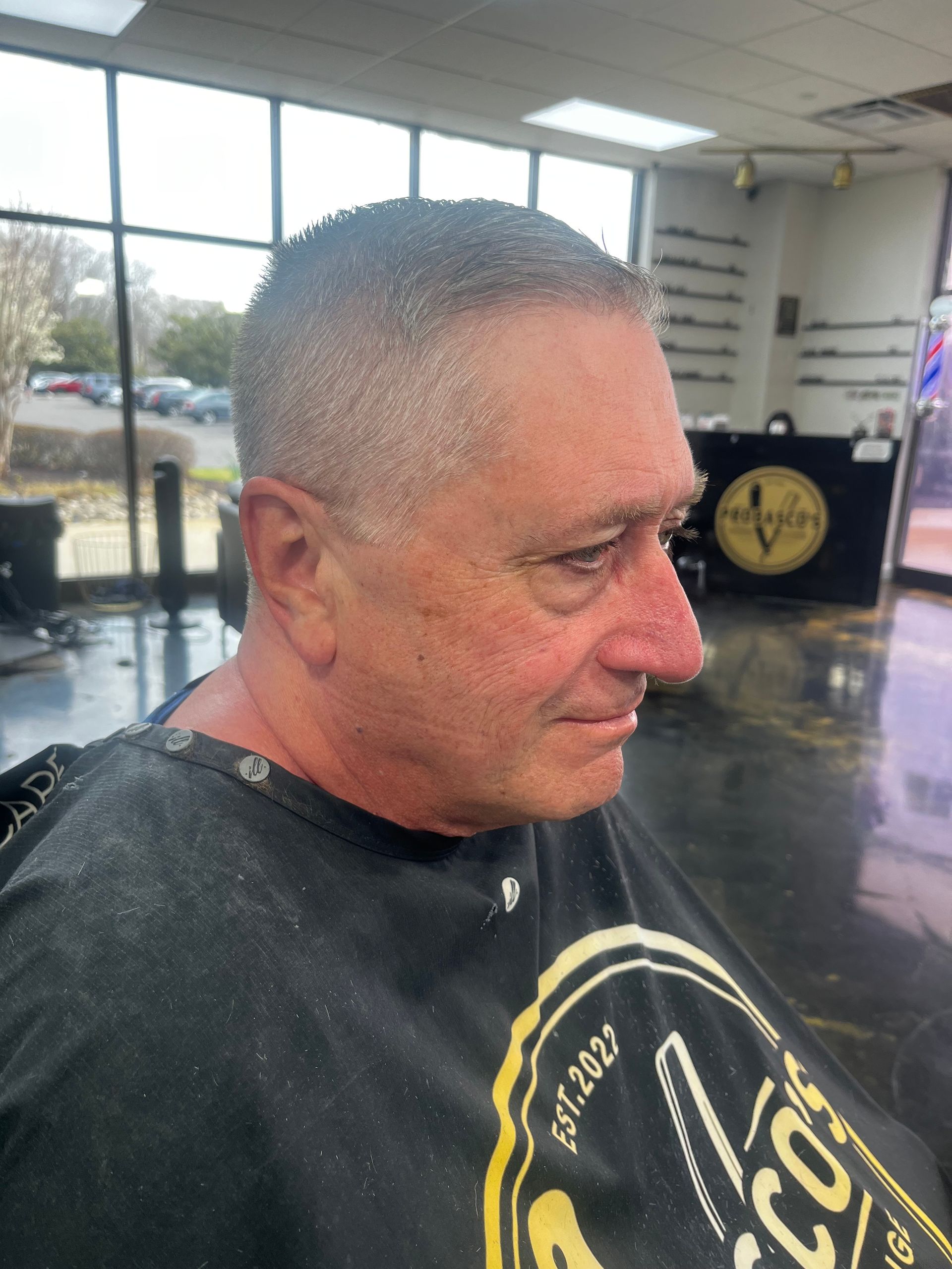 Man in a barber chair with a close-cropped haircut, wearing a black cape in a bright salon.