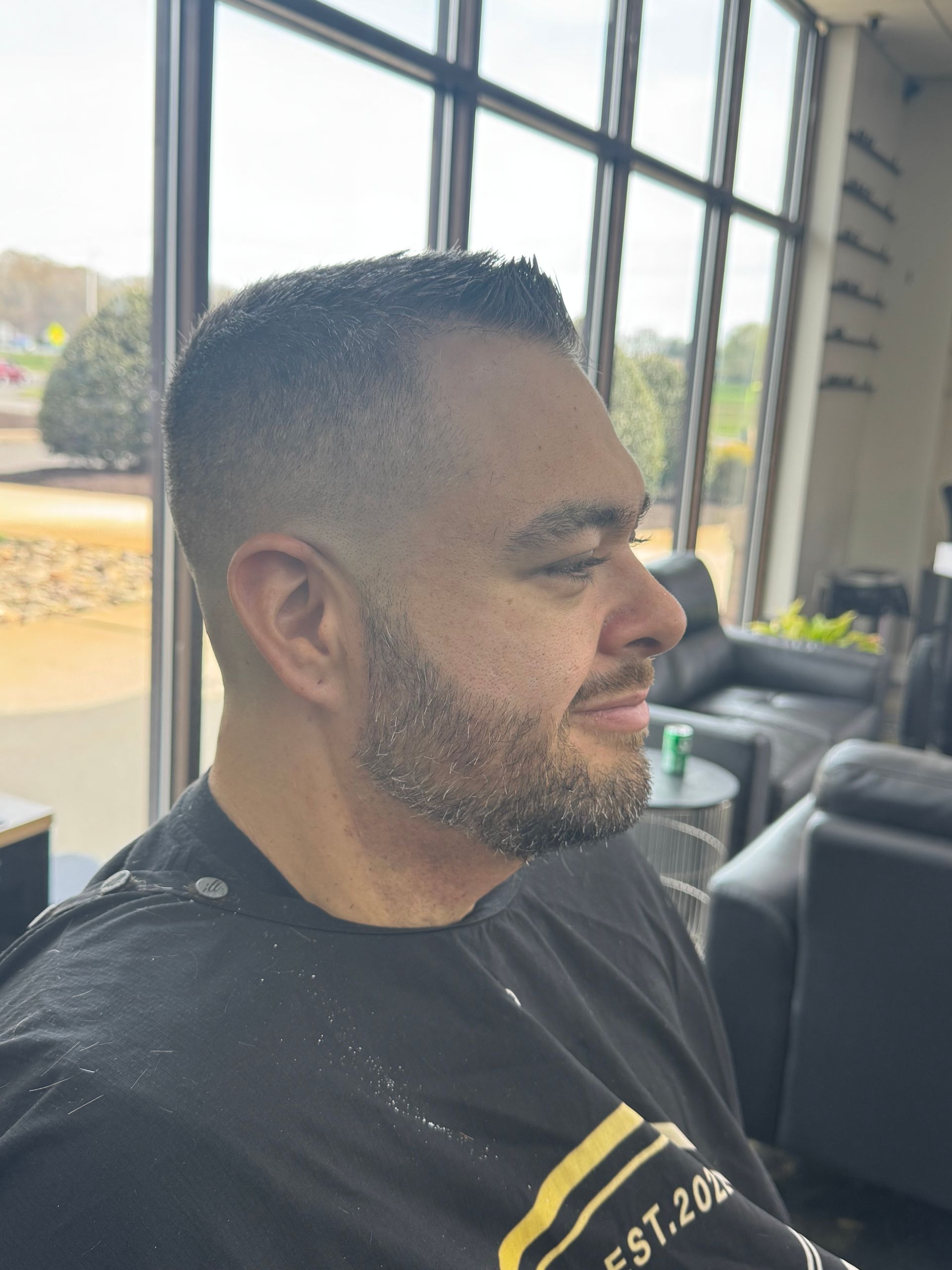 Man in a black T-shirt sitting in profile by large windows, with a trimmed beard and short fade haircut.