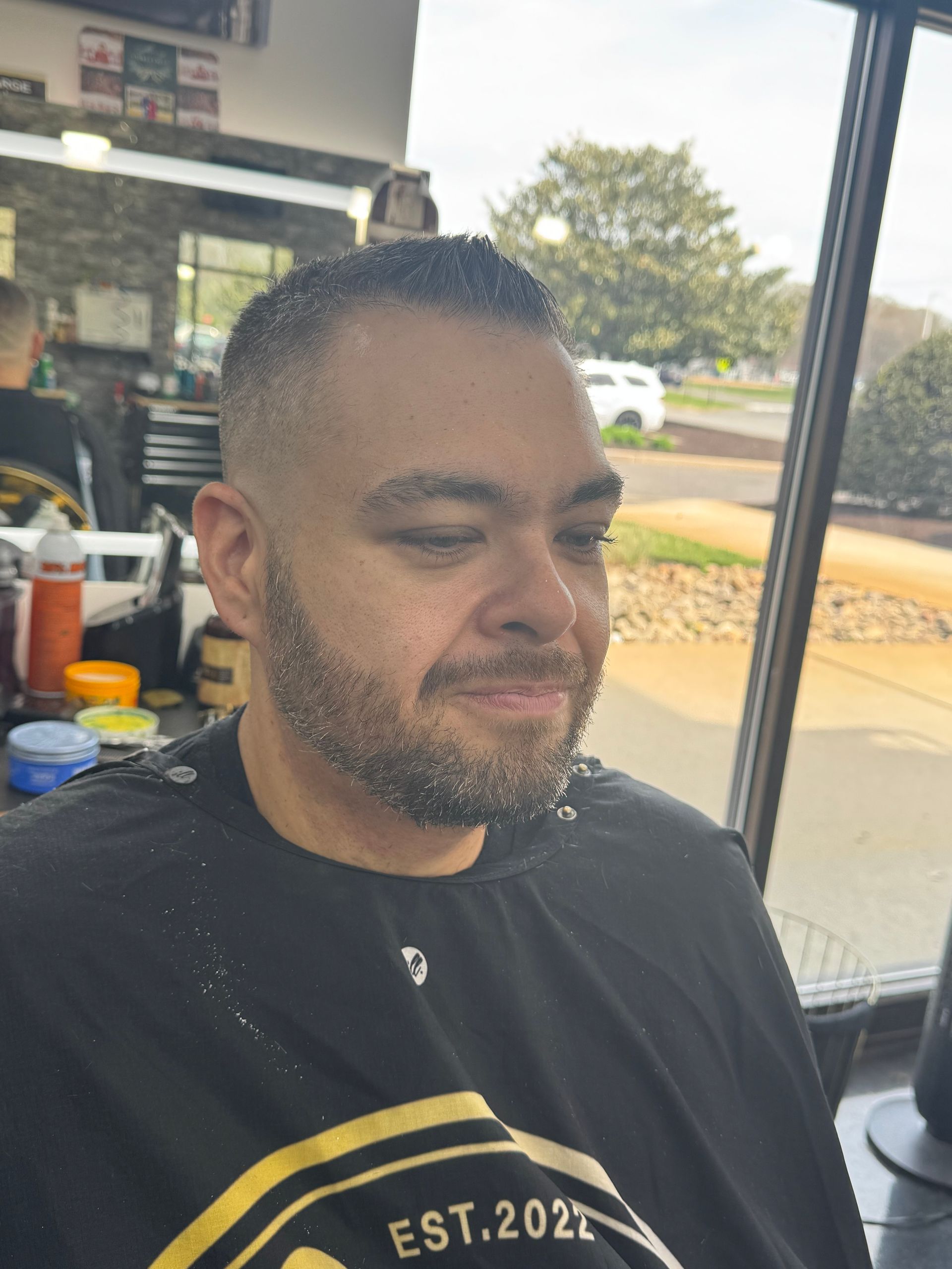 Man sitting in a barbershop chair, wearing a black shirt, with a trimmed beard and a neutral expression.