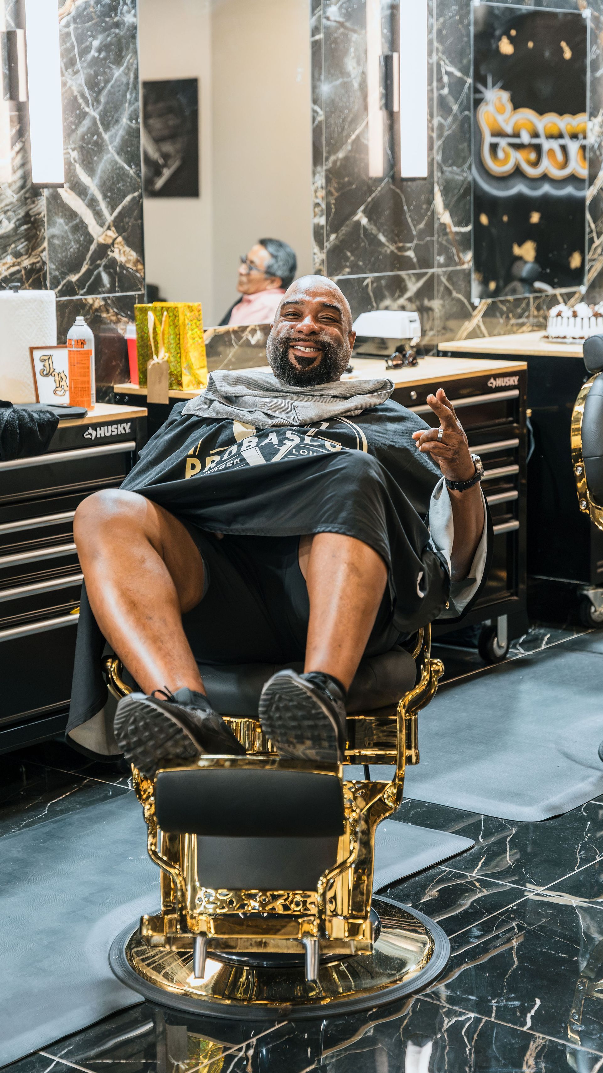 Man reclining in a luxury barber chair in a marble-floored salon, smiling with legs crossed.