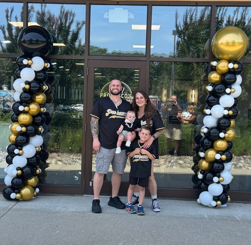 Family posing between black, white, and gold balloon columns outside a building entrance