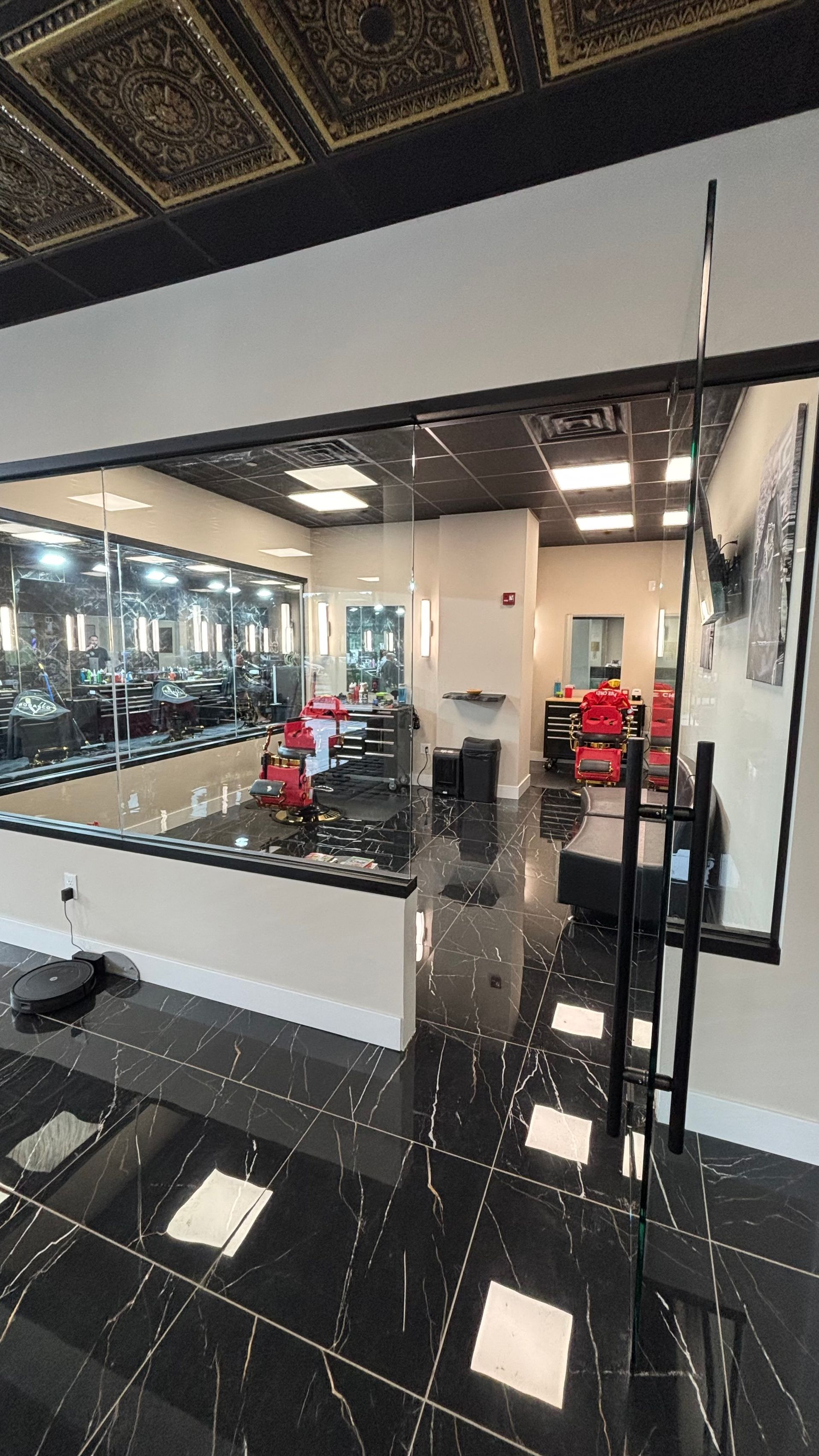 Modern barbershop interior with glossy black marble‑style flooring, red barber chairs, mirrors, and bright recessed lighting, separated by a glass wall and door.