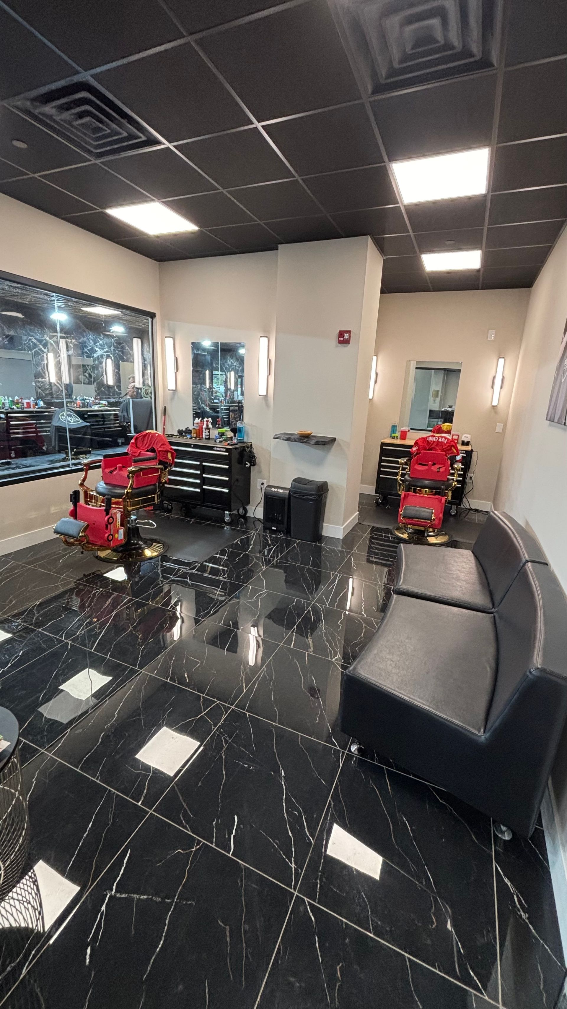 Modern barber shop interior with black tile floor, red styling chairs, mirrors, and black waiting chairs